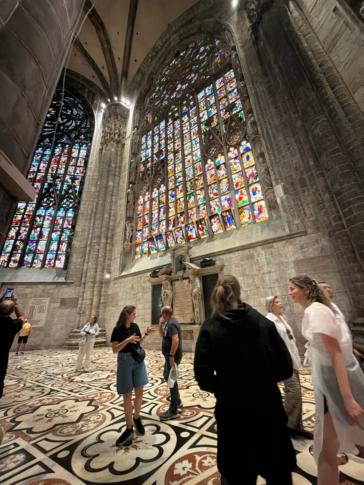 The image depicts the interior of the Milan Cathedral, also known as the Duomo di Milano, in Milan, Italy. The scene is dominated by the cathedral's impressive Gothic architecture, characterized by tall stone pillars, vaulted ceilings, and large, ornate stained-glass windows. Sunlight streams through the windows, illuminating the interior with colorful light.
In the foreground, several people are visible. A woman in the center is wearing a black t-shirt and denim shorts, with a fanny pack around her waist. She appears to be gesturing and talking to a man to her right, who is wearing a dark t-shirt and jeans and holding a piece of paper. To the right of this pair, a woman in a white dress is interacting with them, her right arm raised. Further to the left, a man is taking a picture with his phone, and behind him, a woman in white pants and a light blue denim jacket is standing. In the background, more individuals can be seen observing the cathedral's architecture and stained glass.
The floor of the cathedral is covered in a highly decorative tiled pattern, featuring intricate geometric designs and floral motifs in shades of brown, beige, and white. The overall atmosphere is one of awe and wonder, as visitors explore the vast and ornate space of the cathedral. The image captures the scale, beauty, and spiritual significance of this iconic Milanese landmark.