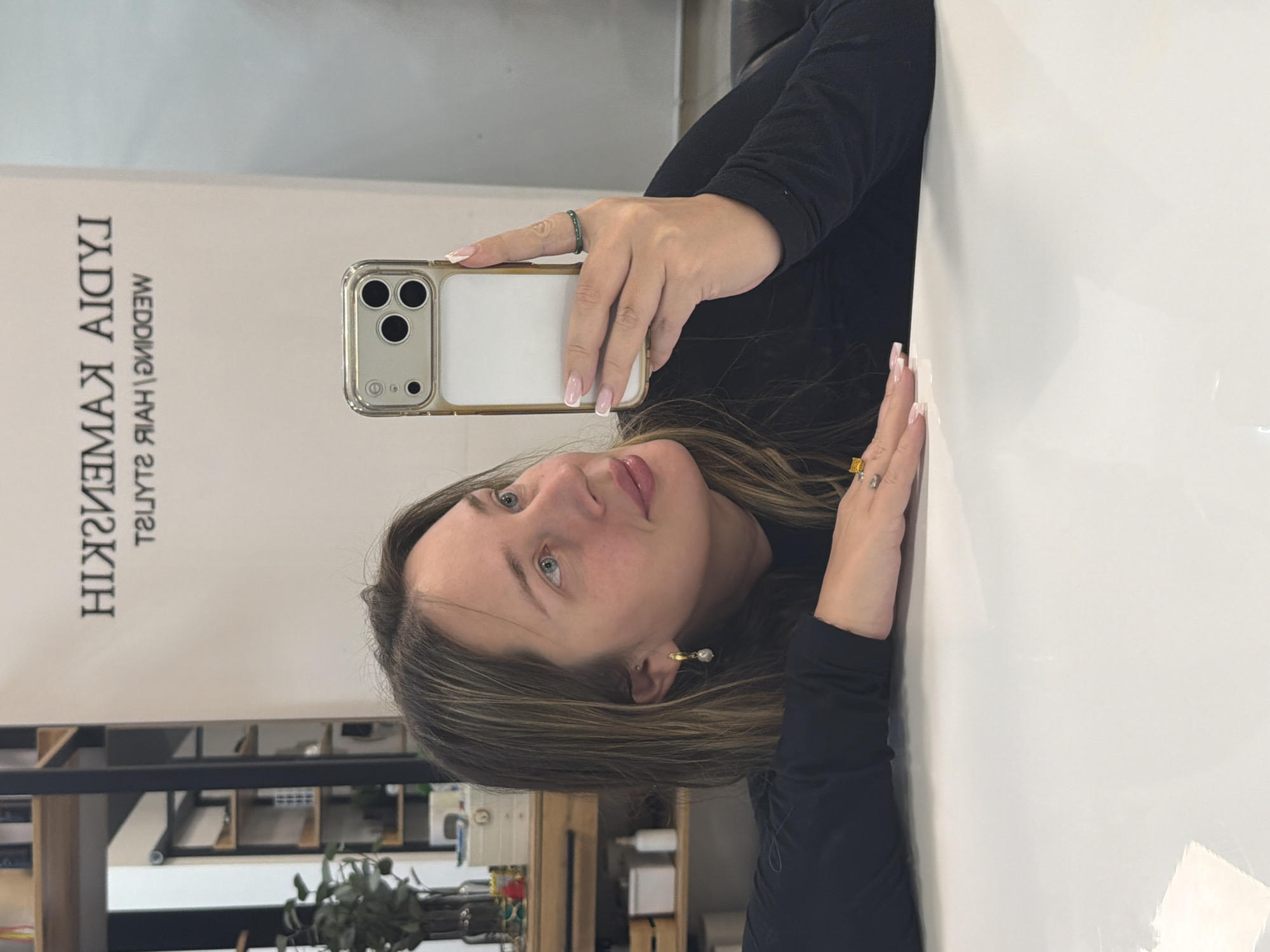 A woman is taking a selfie in a room with a sign that says "Lydia Kamenskiy Wedding Hair Stylist" in Russian. The phone is a gold-cased iPhone with a white back. The woman is wearing a black long-sleeved shirt. Her hair is brown with blonde highlights, and she is wearing pearl earrings and rings on her fingers. The room has a shelf in the background with various items on it. Based on the sign, the location is likely in the city of Chaikovsky, Russia.