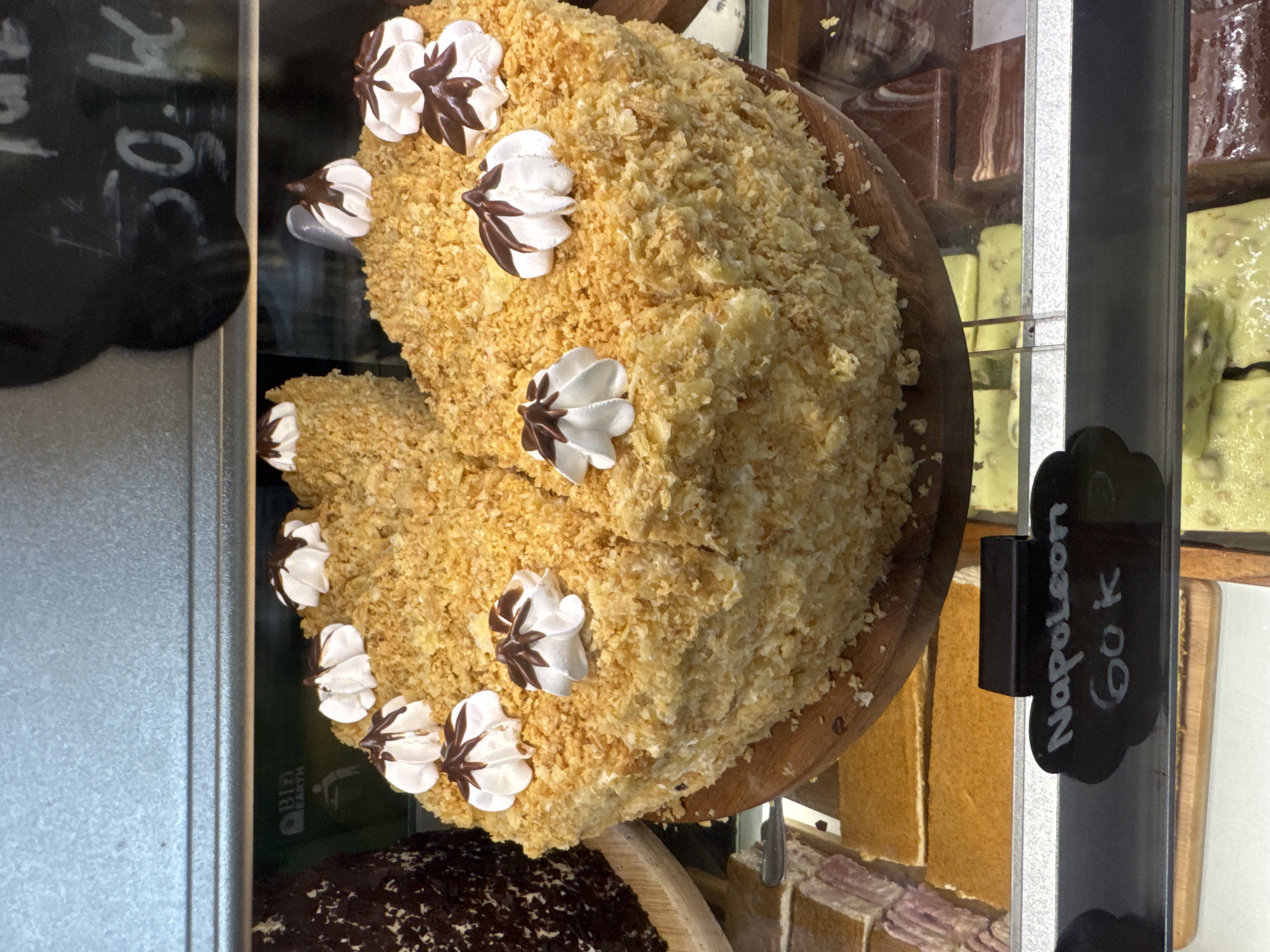 The image shows a Napoleon cake in a bakery display case. The cake is round with a light brown, crumbly surface and decorated with white cream rosettes, some with brown accents. A small black sign next to the cake reads "Napoleon 60k". Other baked goods are visible in the display case. The cake is for sale, with the price listed in Indonesian Rupiah, suggesting the location is likely in Indonesia. The specific location, Kuta Selatan, Indonesia, is inferred from the currency and general bakery context. No people are visible.