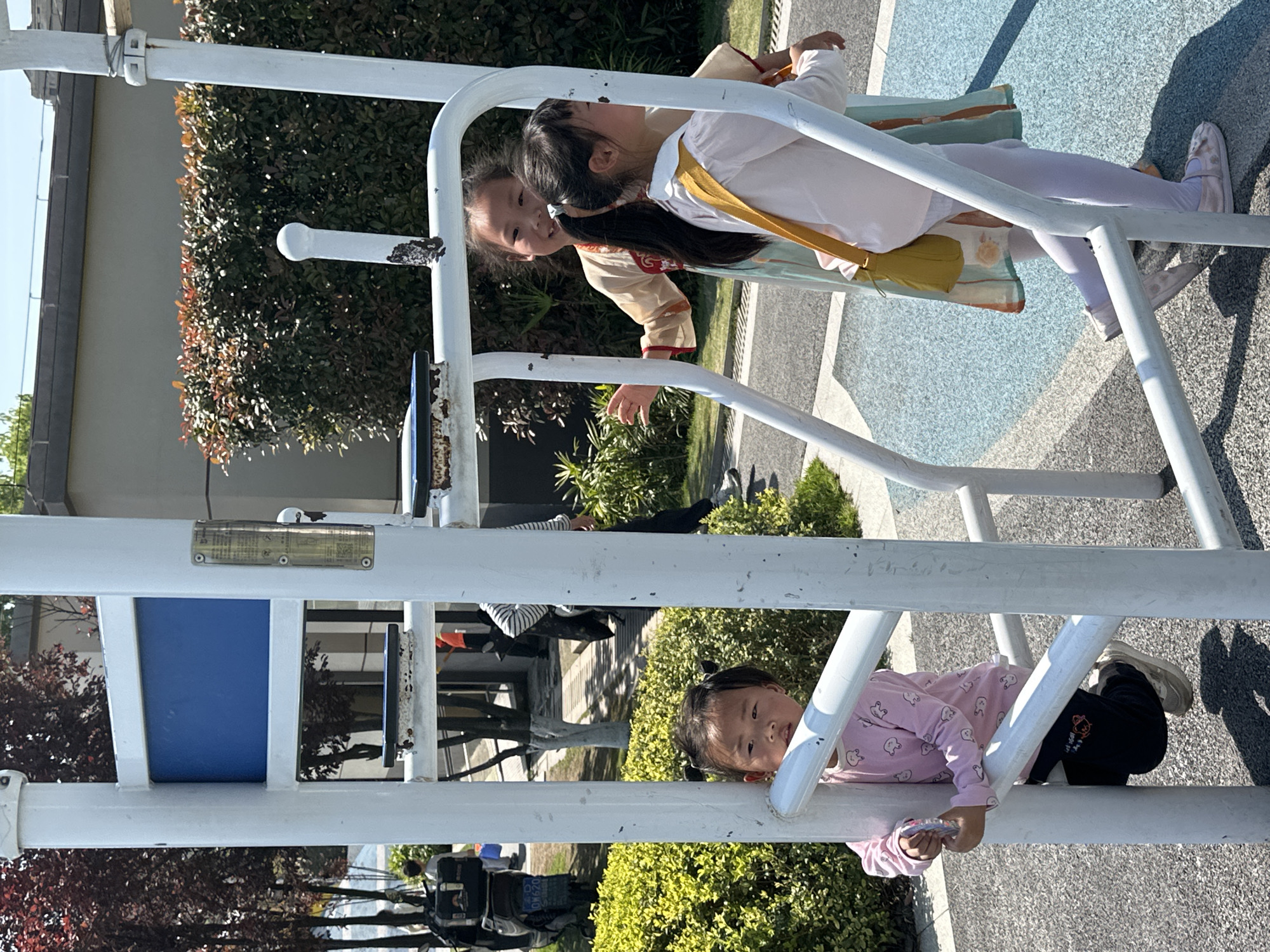 Three young children are playing on a metal climbing frame in an outdoor park. Two girls are positioned near the top of the frame, wearing light-colored clothing, one with a yellow sash and the other in light lavender pants. A third girl is at the bottom of the frame, wearing a light pink shirt with black pants and holding a small card. The frame is white, with blue accents on some sections. The area surrounding the frame includes a concrete pavement and various types of greenery, including bushes and trees. In the background, a street with parked vehicles, including a motorcycle and a car with a license plate number starting with "029" can be seen, and there is a building with an awning. Based on the vehicle license plate, it is likely that this location is in Jiangjin, China.