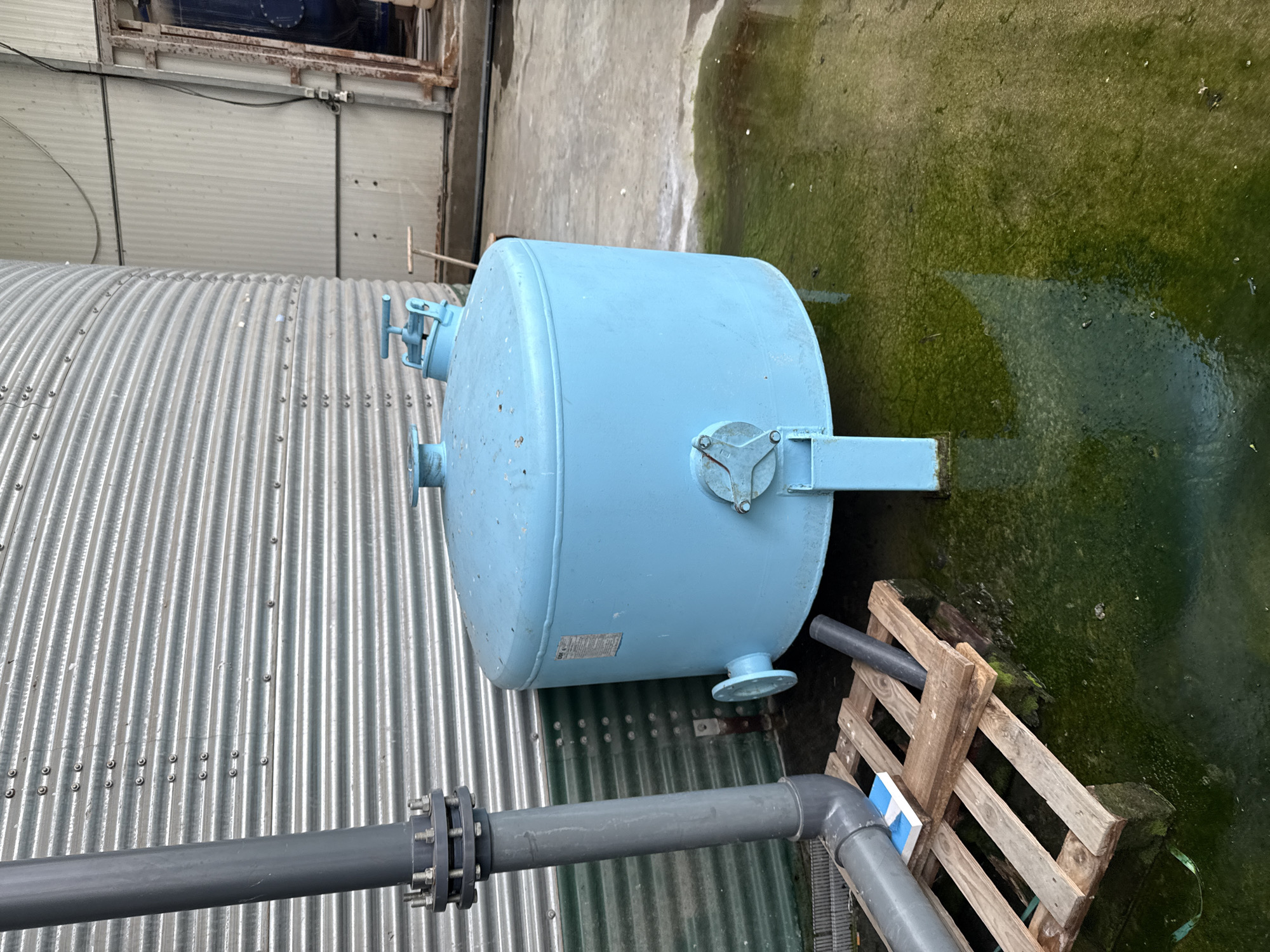 The image depicts an industrial scene. A light blue, cylindrical metal tank sits atop corrugated metal. To the right of the tank is a green, moss-covered concrete surface. A gray pipe with bolted flanges crosses the bottom of the frame, running parallel to the corrugated metal. A wooden pallet is located near the tank's bottom. The location is not identifiable as Stara Zagora, Bulgaria based on the visual elements present.