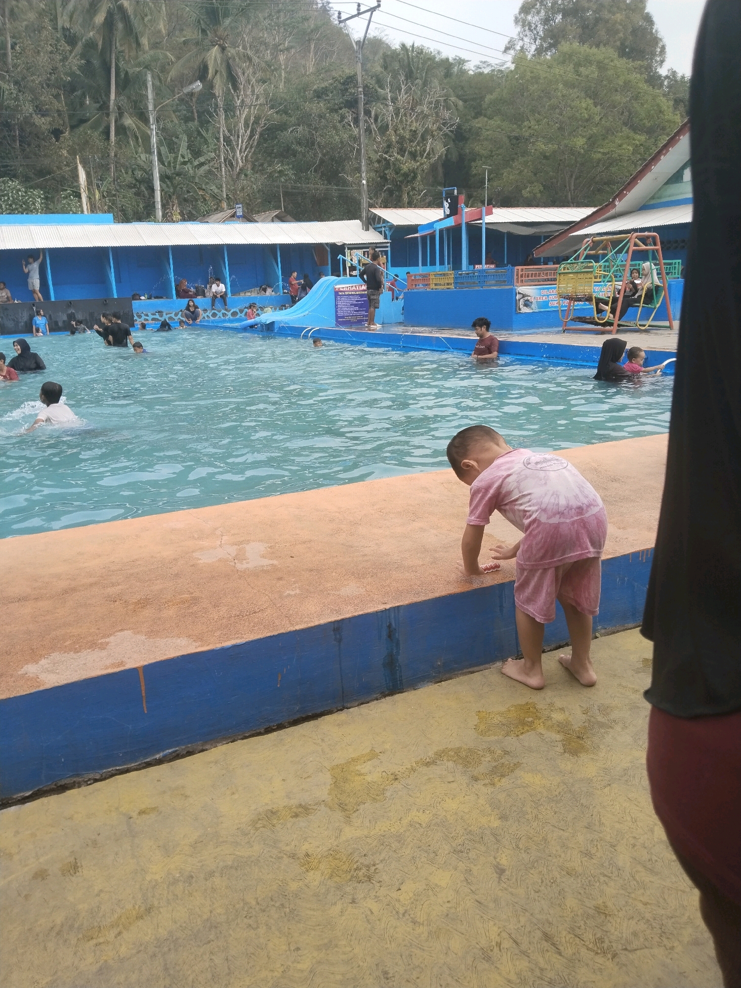 This is a medium shot, eye-level, outdoor photograph taken at a public swimming pool in Batukuwung, Indonesia. The water is a clear blue and appears to be chlorinated. In the foreground, a young boy wearing a pink tie-dye t-shirt and shorts stands on the pool's edge, looking down at his feet. The pool's edge is made of concrete with an orange-brown color on top and a blue painted vertical section below. Behind the boy, the pool is filled with various people of different ages swimming and wading. Some are seen in the background near a blue building with a white roof. To the right, partially visible, is a person wearing a black headscarf and red shorts. The background shows lush green trees and a gentle slope leading up a hill. The sky is overcast. The general atmosphere suggests a recreational area on a warm day.