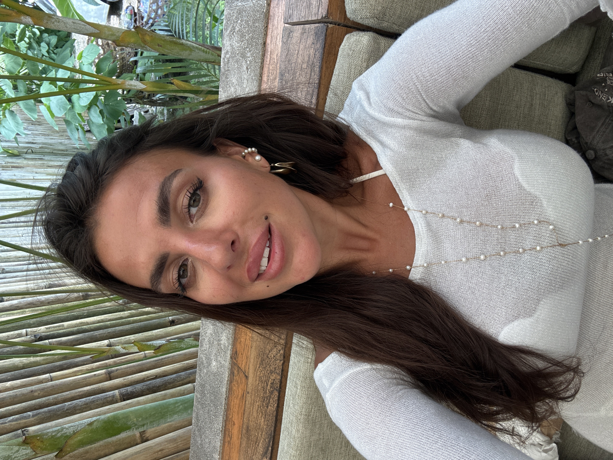 A medium shot shows a woman with long, dark brown hair, wearing a white top and a pearl necklace. She is sitting on a couch, with a bamboo wall and lush green plants in the background. The woman is looking at the camera with a slight smile. A mole is visible on her left cheek, and she is wearing a large hoop earring and a smaller pearl earring in her right ear. The couch appears to be part of an outdoor seating area, possibly at a resort or restaurant in Kuta Selatan, Indonesia. The overall atmosphere is relaxed and tropical.