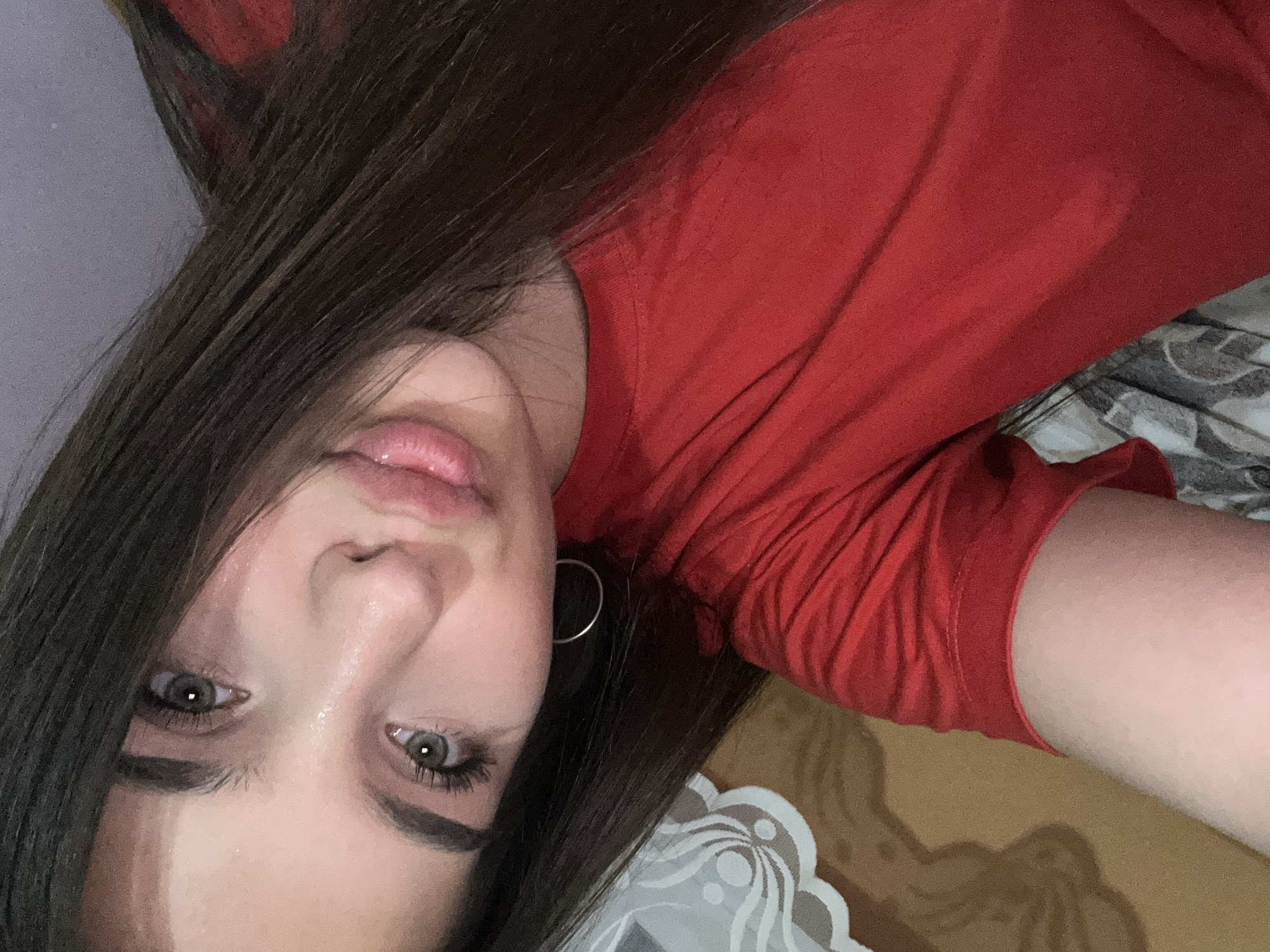 This is a close-up selfie of a young woman with long, dark brown hair. She is wearing a bright red t-shirt. Her face is tilted, and her eyes are looking towards the camera. She has light-colored eyes, defined eyebrows, and full lips. A silver hoop earring is visible in her left ear. The background appears to be an indoor setting with a patterned curtain or fabric on the left and a textured surface, possibly furniture or bedding, behind her. There is no identifiable information that places this image in Shumen, Bulgaria.