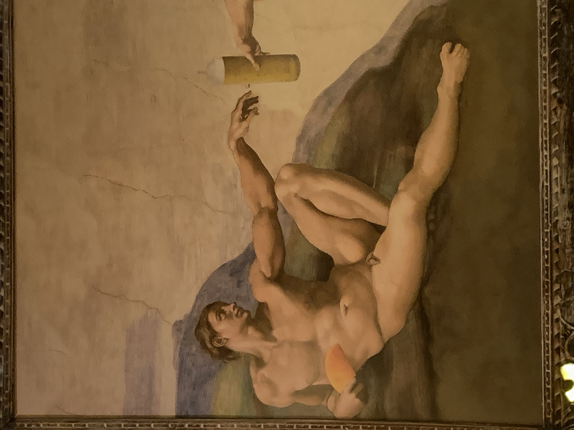 The image depicts a framed painting, likely a parody or modern interpretation of Michelangelo's "The Creation of Adam" from the Sistine Chapel. The figures are similar in pose, but the object being exchanged is a beer can instead of a divine spark. The figure on the right is holding a mango in his left hand. The location is not discernible from the image, and there is no evidence indicating it is in Barcelona, Spain. The environment appears to be a gallery or building with a large collection of art, as indicated by the frame.