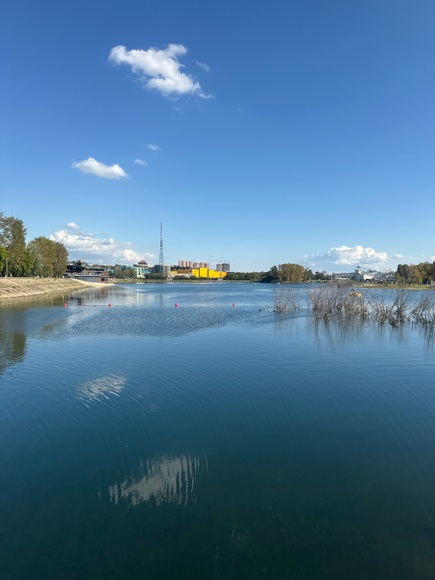 The image depicts a wide, calm body of water, likely a lake or large pond, under a bright blue sky with scattered white clouds. The water's surface reflects the sky and some surrounding vegetation. On the left bank, there is a concrete embankment, lined with trees. Beyond the trees, a cityscape is visible, featuring buildings of varying heights and colors, including a prominent tall radio tower and a large yellow structure. Several red buoys are floating in the water, indicating a designated area. In the middle ground, some bare branches of trees emerge from the water. Further in the distance, on the right side of the water, more trees and some structures are visible. The scene appears to be a public recreational area, possibly a park or waterfront in the city of Irkutsk, Russia. There are no discernible people in action.