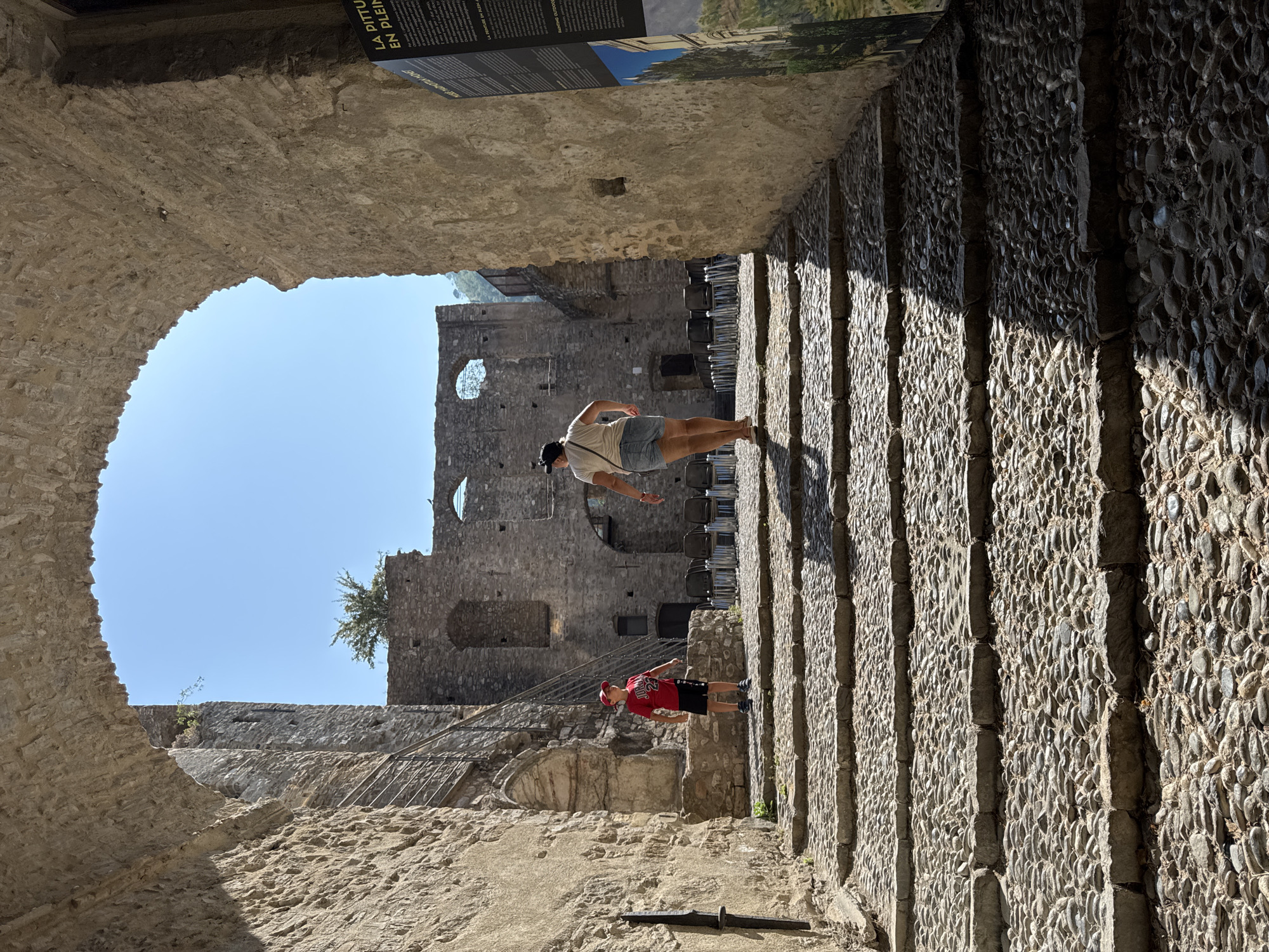 The image captures a view looking through a stone archway towards a cobbled stairway within the medieval village of Dolceacqua, Italy. Two individuals are visible on the stairs. A young boy wearing a red shirt, black shorts, and a red cap stands on a lower step. Further up the stairs, a woman with a light-colored t-shirt and denim shorts walks away from the camera. Behind them, the ruins of a stone structure are visible, with a clear blue sky overhead. A vertical sign with text and an image is positioned on the right side of the frame. The stonework of the archway and the surrounding buildings suggests a historical setting.