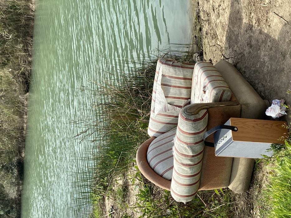 A static outdoor scene depicts a worn armchair and a portable speaker situated on a dry, cracked dirt bank adjacent to a body of water in the city of Unknown, Unknown. The armchair, positioned near the water's edge, features striped upholstery in light beige with red vertical lines on its seat, backrest, and armrolls, exhibiting multiple tears and significant wear. Its outer frame and base cushions are a solid reddish-brown and light beige respectively. To the left of the armchair, a rectangular portable speaker with a light wooden side panel, a light grey control panel with silver knobs, and a dark handle stands upright on the ground. A small crumpled piece of white and colored material lies on the dirt near the speaker. The body of water, occupying the background, is a calm, light teal-green color with subtle surface ripples. The far bank across the water consists of dense green and brown vegetation. Sparse patches of green grass and tall dry reeds grow along the immediate bank. The scene is brightly illuminated by natural daylight. No individuals are present.