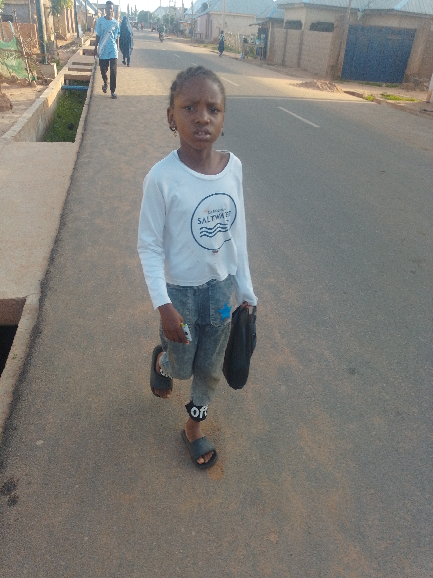 A young girl with braided hair and hoop earrings is captured in a medium shot, walking on a paved street in a city. She is wearing a white long-sleeved shirt with a graphic print that reads "CAROLINA SALTWATER" and denim jeans with a blue star patch on the back pocket. She is also wearing dark-colored slip-on sandals and has a black bag in her right hand. To her left, a shallow concrete drainage ditch runs alongside the street. In the background, a man in a light blue t-shirt and black pants is walking away from the camera, and a woman wearing a dark headscarf and abaya is also present. Other individuals are visible in the distance, along with buildings and a parked vehicle. The scene appears to be during daylight hours.
