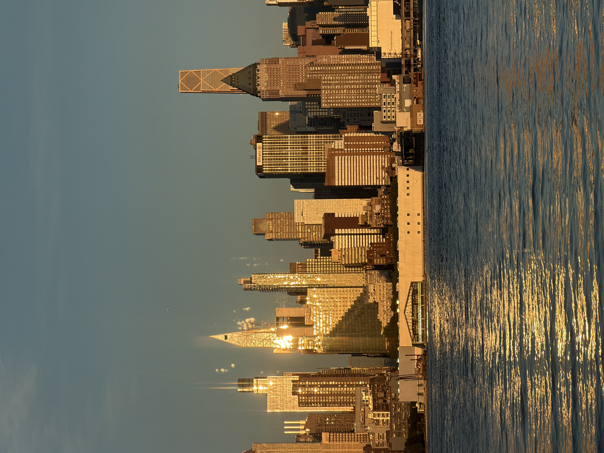 A panoramic view of the New York City skyline is captured from Weehawken, New Jersey, at golden hour. The sun's warm, low light glints off the glass facades of numerous skyscrapers, creating a dazzling spectacle. In the foreground, the calm waters of the Hudson River reflect the vibrant colors of the sunset and the city lights. A large, modern building with a glass-fronted interior and a peaked roof stands prominently along the waterfront. Among the towering structures, an airplane can be seen flying in the distance, a small speck against the vast expanse of the sky. The image depicts a serene and majestic urban landscape at the close of day.