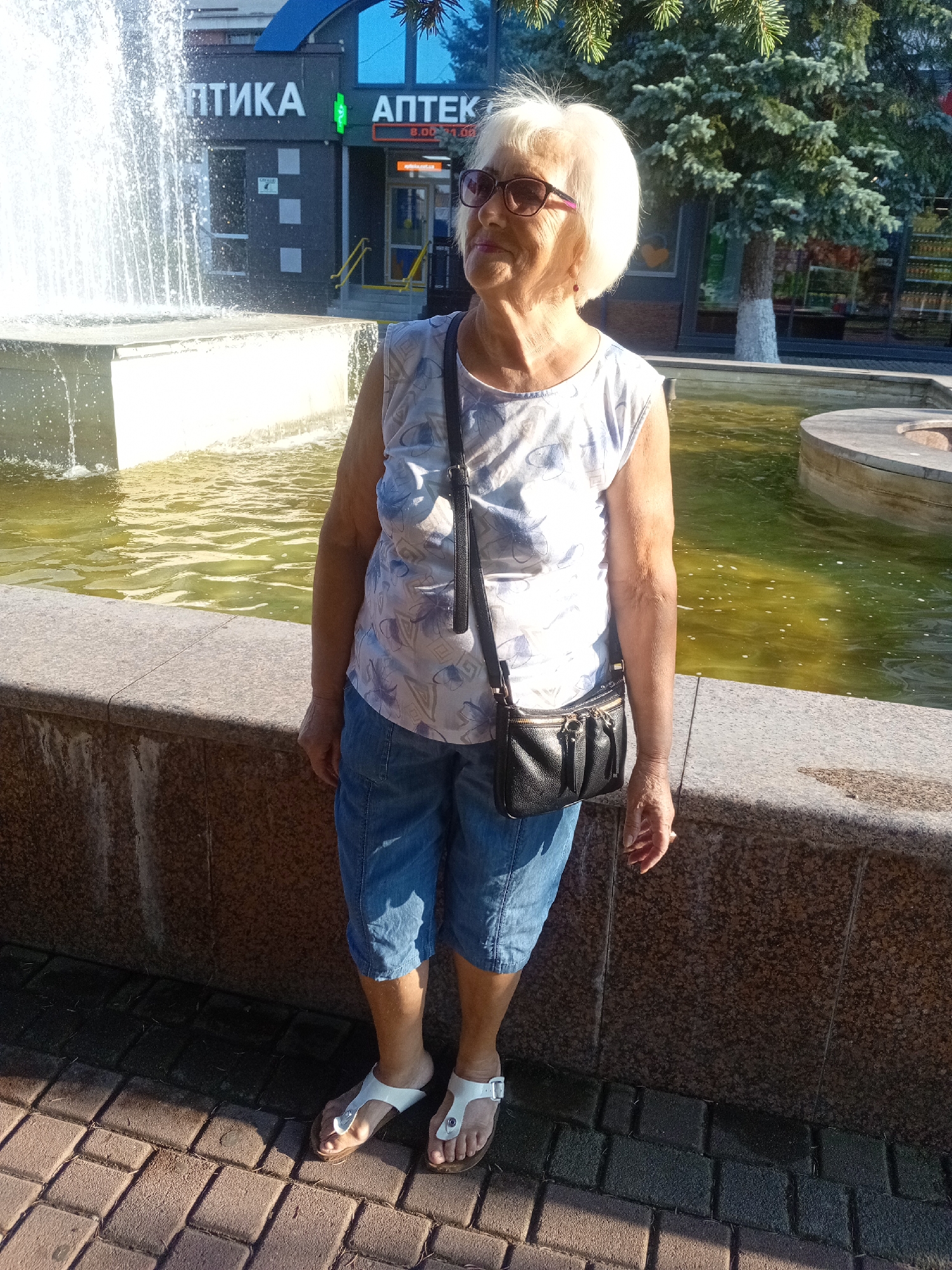 An elderly Caucasian woman with blonde hair and sunglasses stands by a fountain in an urban setting, likely Tiachiv, Ukraine, as suggested by the Cyrillic lettering on a building that reads "АПТЕКА" (Pharmacy) and "ОПТИКА" (Optics). She is wearing a sleeveless white shirt with a blue floral pattern and denim capri pants, along with white sandals. A black leather crossbody bag with gold zippers is slung over her shoulder. The fountain water is murky green, and the surrounding area is paved with brick-like stones. A large evergreen tree is visible behind the woman, and a building with windows displaying products is in the background. The lighting suggests it is daytime, with sunlight casting shadows on the pavement.