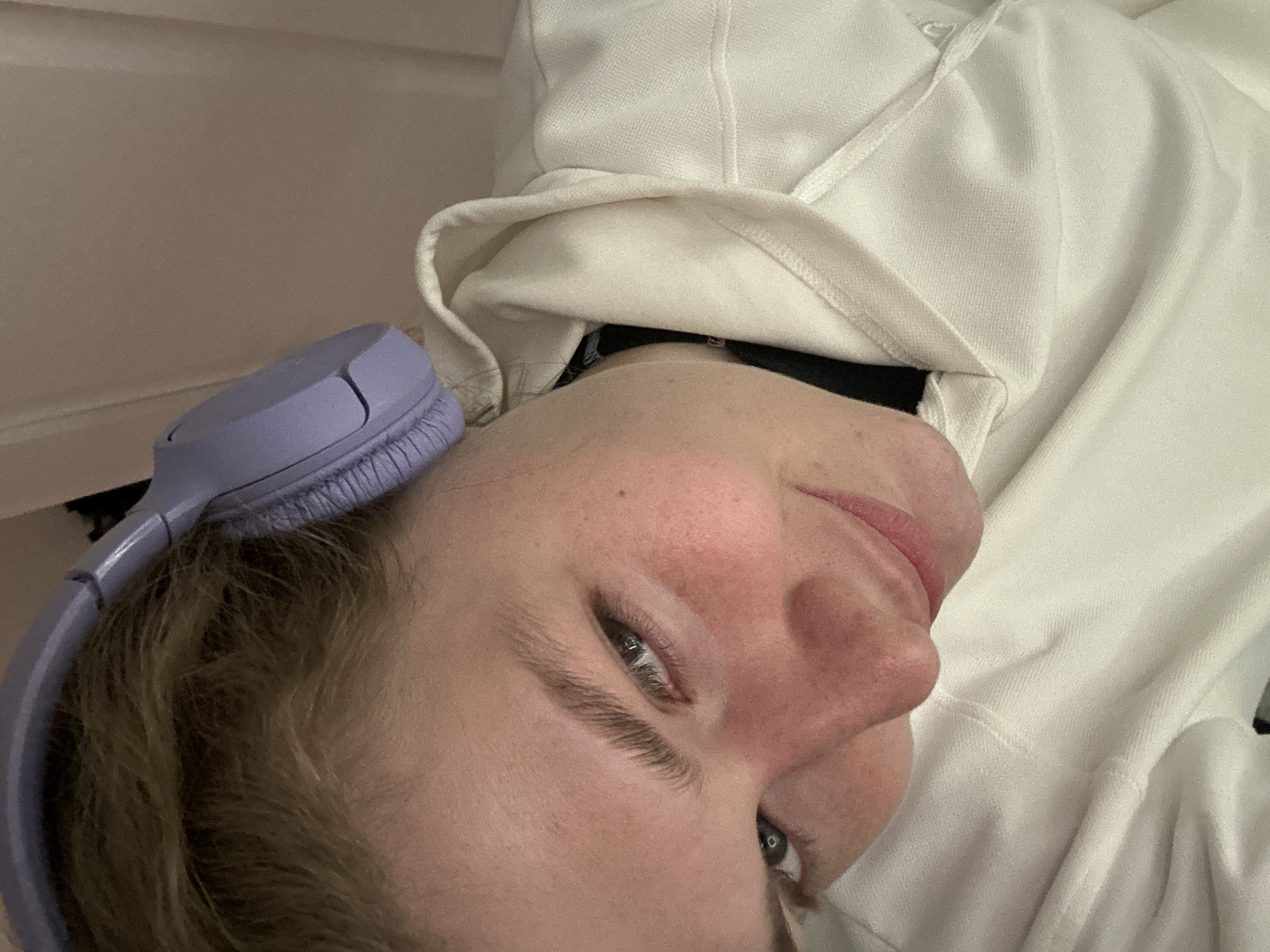 A close-up, rotated image shows a person wearing lilac headphones resting on their head. The person has fair skin with pink cheeks, light eyebrows, and visible eyelashes. Their left eye is partially visible and blue-grey. They wear a white hooded sweatshirt. A dark object, possibly clothing, is visible beneath the hood. The background is a pale neutral color. The location is not discernible from the image.