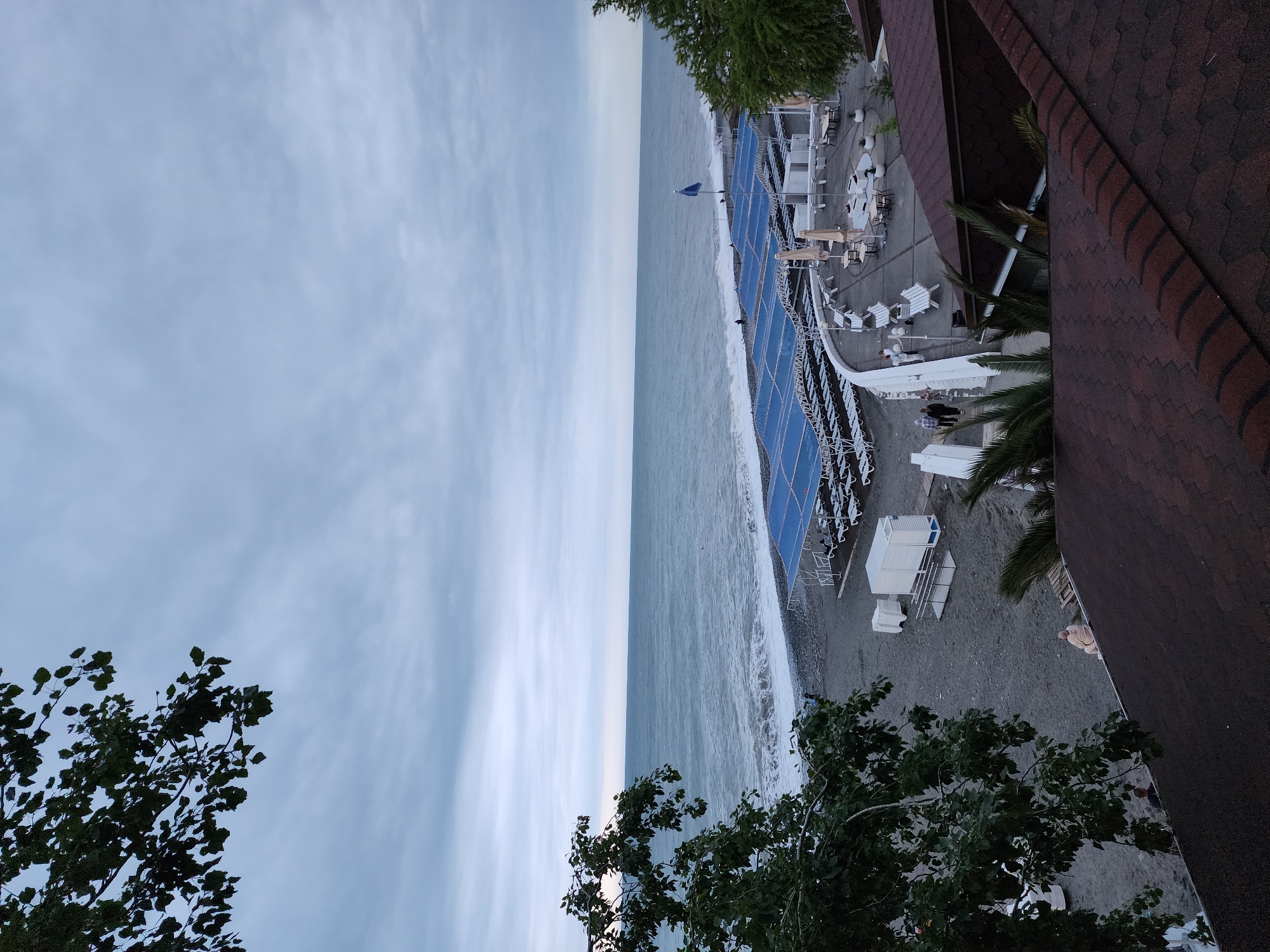 This image shows an elevated view of a beach area, likely in Sochi, Russia. The sky is overcast with visible cloud formations. The sea is a muted blue-grey, with small waves reaching the pebble-covered beach.
A structure with blue canopies and white support beams runs along the beach, featuring numerous white lounge chairs, tables, and umbrellas, suggesting a private or commercial beach club. Further back, there are smaller, white, building-like structures.
The image is taken from a height, possibly from a balcony or high window, as indicated by the tiled roofing and some leafy trees/bushes visible in the foreground. The foliage frames parts of the bottom and right of the picture. The lack of vibrant colors and the cloudy sky give the scene a calm, somewhat muted aesthetic. There are some people near the structures at the beach.