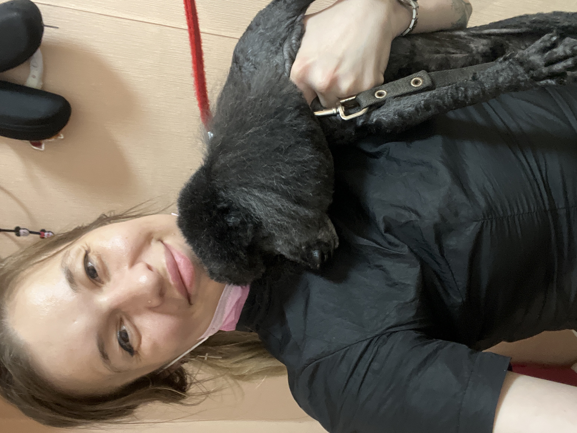 A young woman with light brown hair pulled back, wearing a black, long-sleeved collared shirt and a pink face mask, holds a black poodle puppy. The woman is smiling and looking towards the camera, with a silver nose ring and a cross earring visible. The puppy is resting its head on the woman's shoulder, with its eyes closed. A red leash is visible behind the woman. The background appears to be an indoor wall, possibly in a grooming salon or pet-friendly establishment.