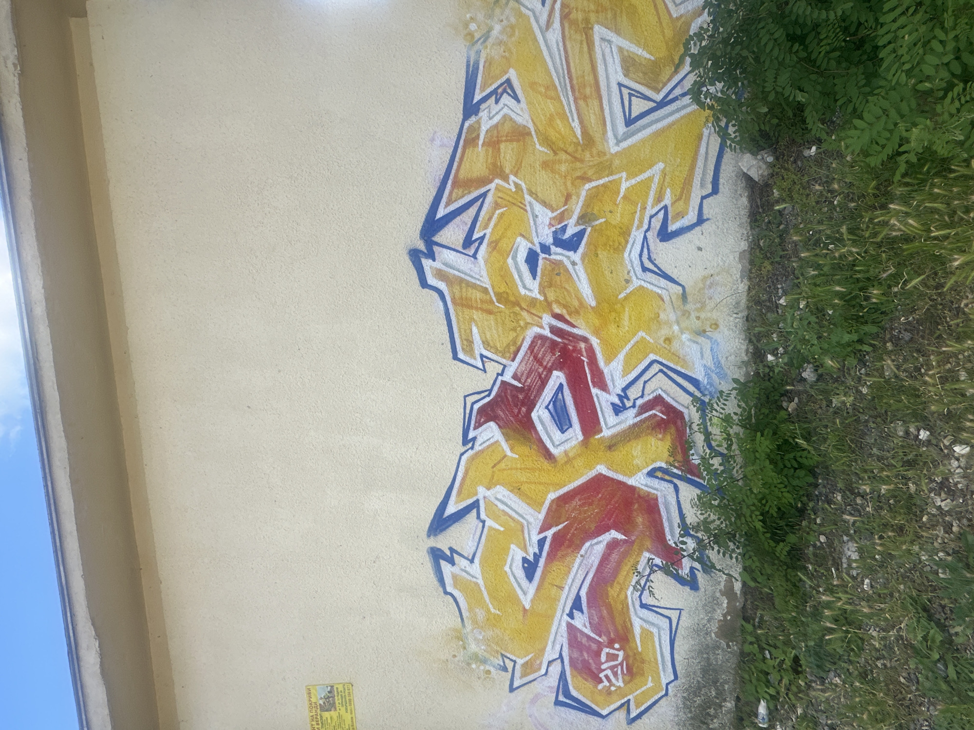 A bright yellow and red graffiti piece adorns a textured, cream-colored wall. The graffiti, with bold blue outlines and white accents, appears to be a stylized word. The letters are sharp and angular, with a layered effect. To the left of the graffiti, a yellow and black advertisement is affixed to the wall. Below the wall, a strip of green foliage and dry grass with scattered small stones covers the ground. The sky above is a clear blue with a few wispy clouds.