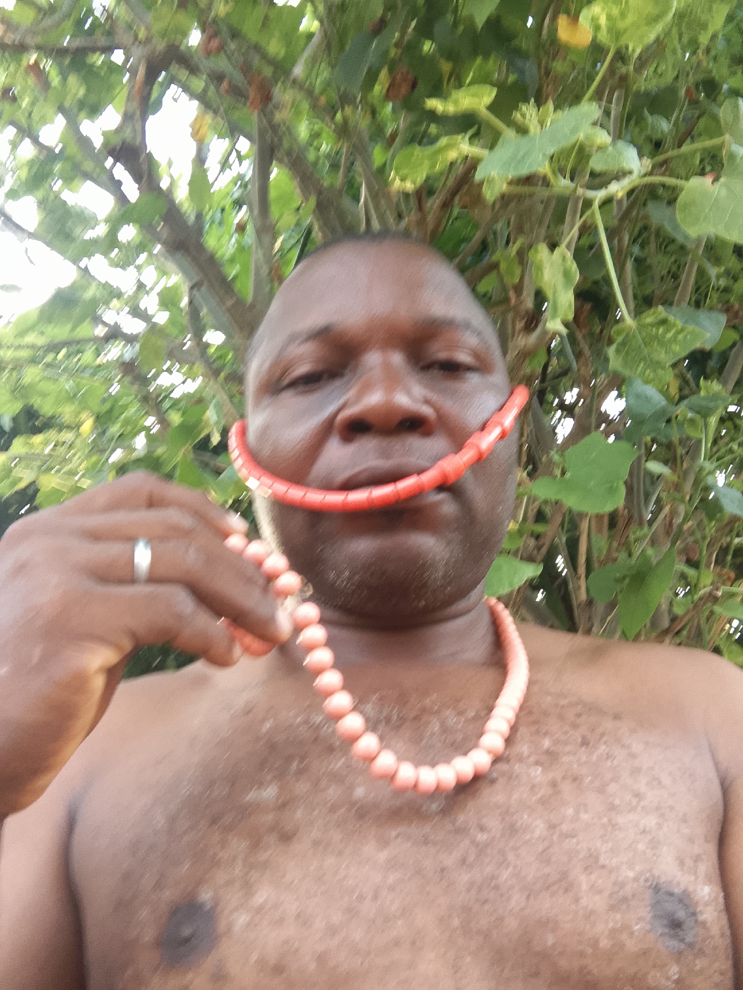 The image shows a dark-skinned, bare-chested man holding a red beaded necklace. A portion of the necklace is positioned in his mouth, mimicking a smile. He is wearing a ring on his left hand. The background is blurred, but appears to be foliage. The location is identified as Unknown, Unknown.