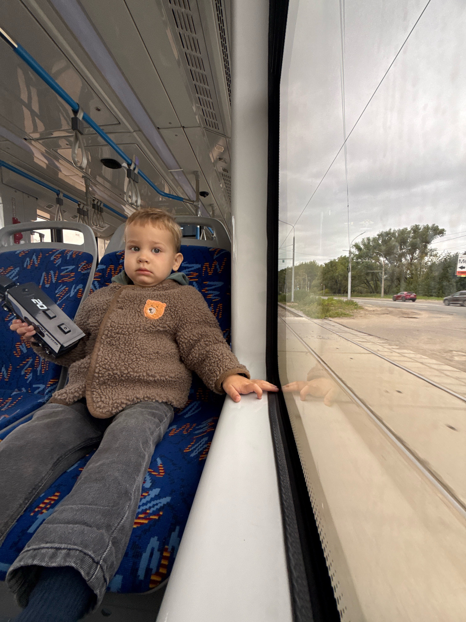 A young Caucasian boy with blonde hair is sitting on a blue and orange patterned seat inside a vehicle, likely a tram. He is wearing a brown fuzzy jacket with a bear patch on the left chest and dark grey pants. In his left hand, he is holding a black electronic device with a digital display showing "20". He is looking directly at the camera with a neutral expression. The interior of the vehicle shows overhead blue support bars and ventilation grates. Through the large window beside him, a street scene is visible with trees, a road, two cars, and tram tracks. The sky is overcast. The location is identified as Rusanovo, Russia.