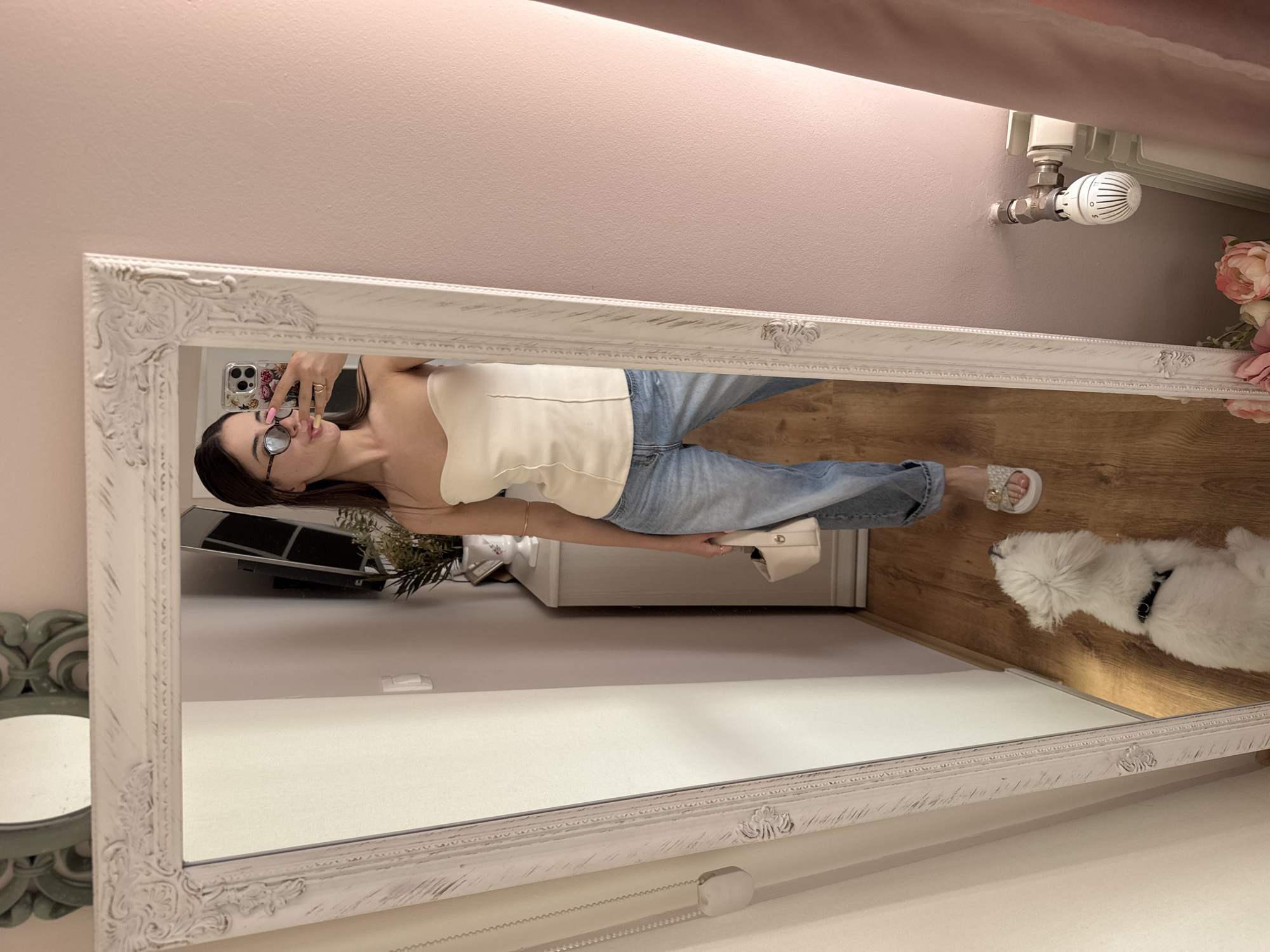 The image captures an indoor scene, likely a room in a home in Targovishte, Bulgaria, with a woman taking a mirror selfie. The main subject, reflected in a large, ornate white mirror with a distressed finish, is a woman with long dark hair wearing glasses, a light-colored strapless top, blue jeans, and white slide sandals. She is holding a smartphone with a floral patterned case to take the picture, and a small white handbag in her other hand. A ring is visible on her right hand.
To the right of the woman's reflection, on the wooden floor, a small white fluffy dog is lying down. The wall behind the mirror is a muted pastel pink or purple. Above the mirror, on the right side of the frame, a white radiator is visible with a thermostatic valve that shows numbers from "0" to "5" and a snowflake icon. Next to the radiator are artificial pink and white peony flowers.
On the left side, another smaller, oval mirror with a green ornate frame is partially visible. Further into the reflection, a dark monitor or TV screen and a green potted plant can be seen. The floor throughout the room is made of light-colored wooden planks. The woman's expression is casual, with slightly pursed lips.