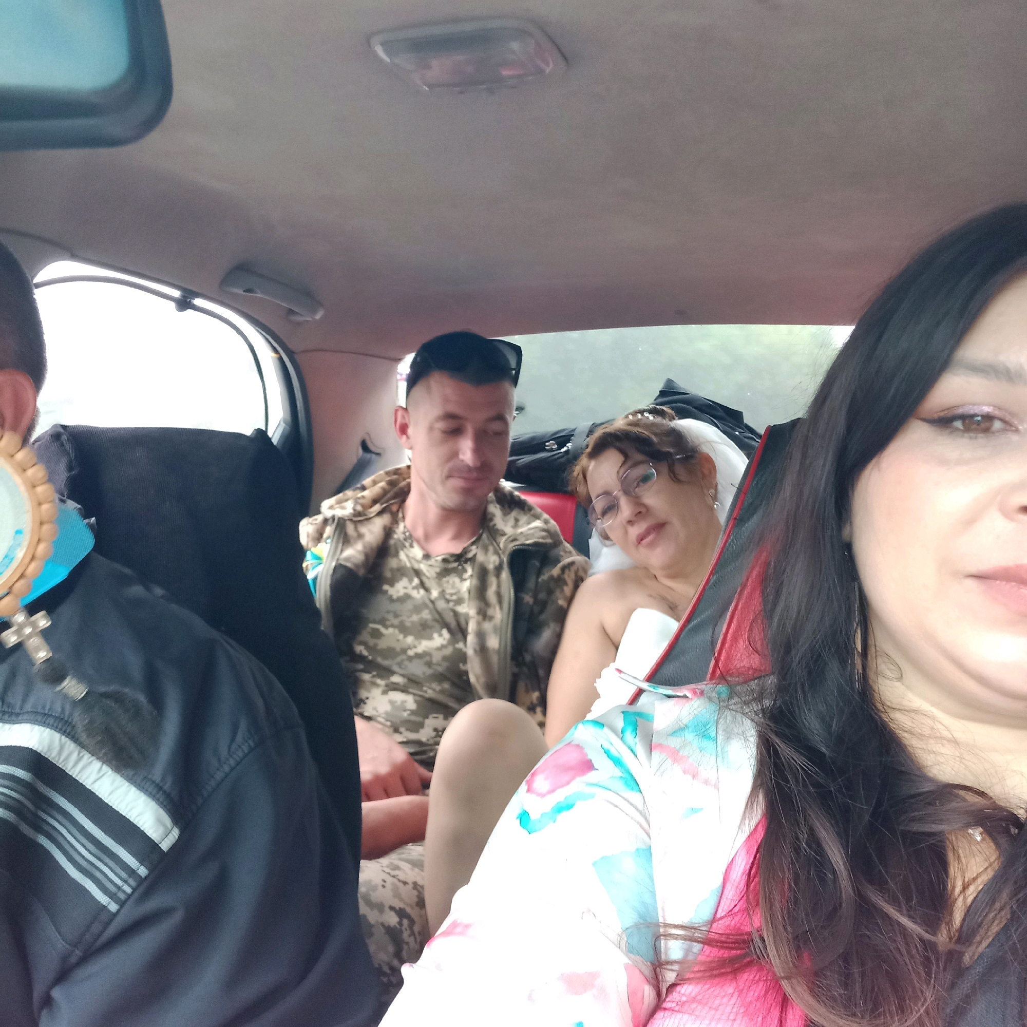 In the interior of a vehicle, four individuals are visible. The leftmost person is partially obscured, wearing a dark jacket with striped detailing on the sleeve. They have a necklace with a cross and a circular pendant. To their right, a man in a camouflage jacket is seated, looking downwards. Further to the right, a woman with glasses is reclining, her upper body bare, and she is holding a white sheet or blanket. In the foreground on the right, a woman with long, dark hair is partially visible, looking towards the camera. The vehicle's interior includes a light-colored headliner and a partially open sunroof. The overall scene suggests travel or a casual gathering within a car.