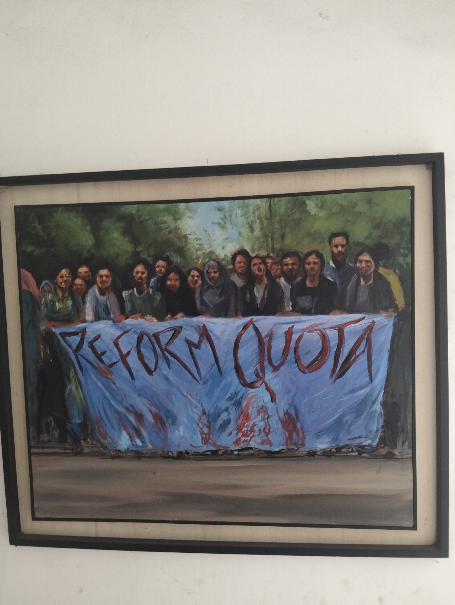 The image shows a framed painting depicting a group of people holding a banner that reads "REFORM QUOTA." The banner is blue with red lettering and apparent red stains below the text. The people appear to be of South Asian descent. The background consists of trees, implying an outdoor setting. The painting is framed and is against a white wall. There are no obvious features to establish the location as Dhaka, Bangladesh, or any city in particular.