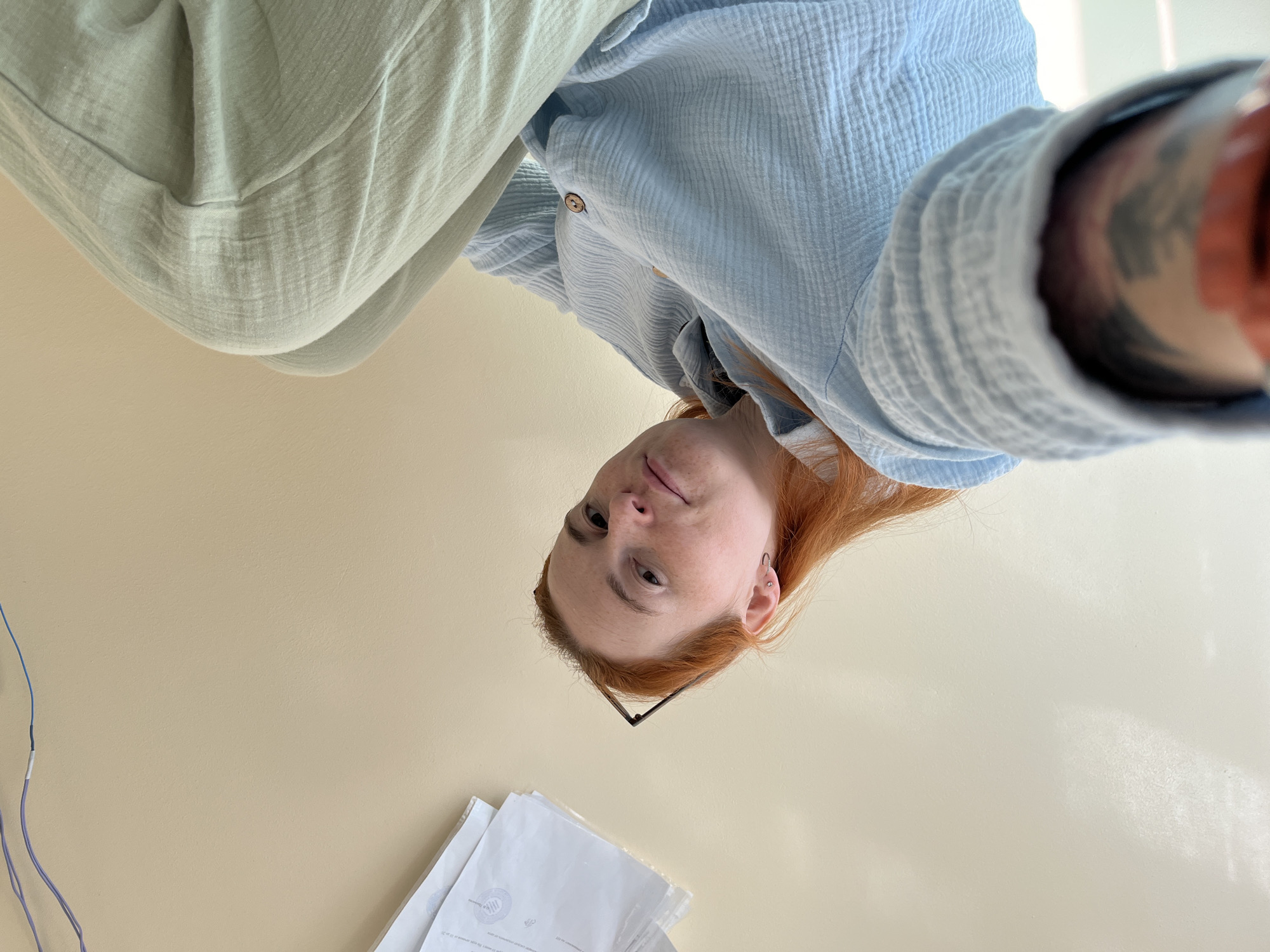 A young woman with red hair and freckles is sitting, looking up at the camera. She is wearing a light blue, crinkled fabric shirt with a wooden button and light green pants. A pair of glasses rests on top of her head. Her left arm is visible, showing a tattoo on her forearm. In the upper right corner, there are some papers and wires attached to the wall. The background is a plain, light-colored wall. The photo is taken from a low angle, looking up at the woman. The location is not identifiable as being in Chaikovsky, Russia.