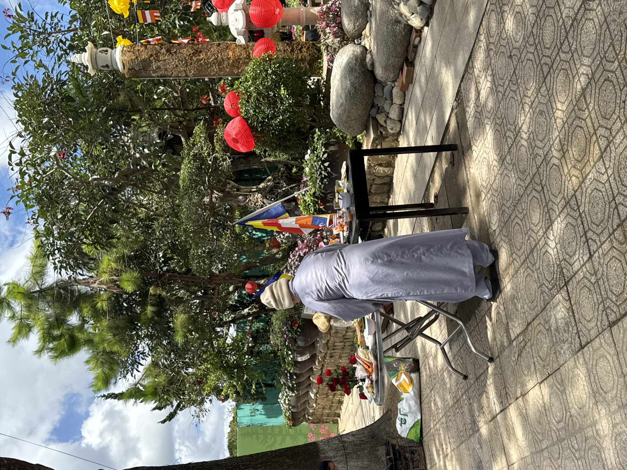 A person dressed in a grey robe and a white beanie is standing with their back to the camera, facing a table laden with food offerings. The scene appears to be outdoors, possibly in the garden of a Buddhist temple or monastery in Da Lat, Vietnam, given the presence of lush vegetation, stone pathways, and decorative lanterns. A flagpole with a Buddhist flag stands to the left of the table. Red lanterns are hung from trees and a stone pillar in the background, adding to the festive or ceremonial atmosphere. The ground is paved with patterned tiles, and large rocks are scattered in the landscaping. A bag of what appears to be rice is on the ground near a metal folding table.