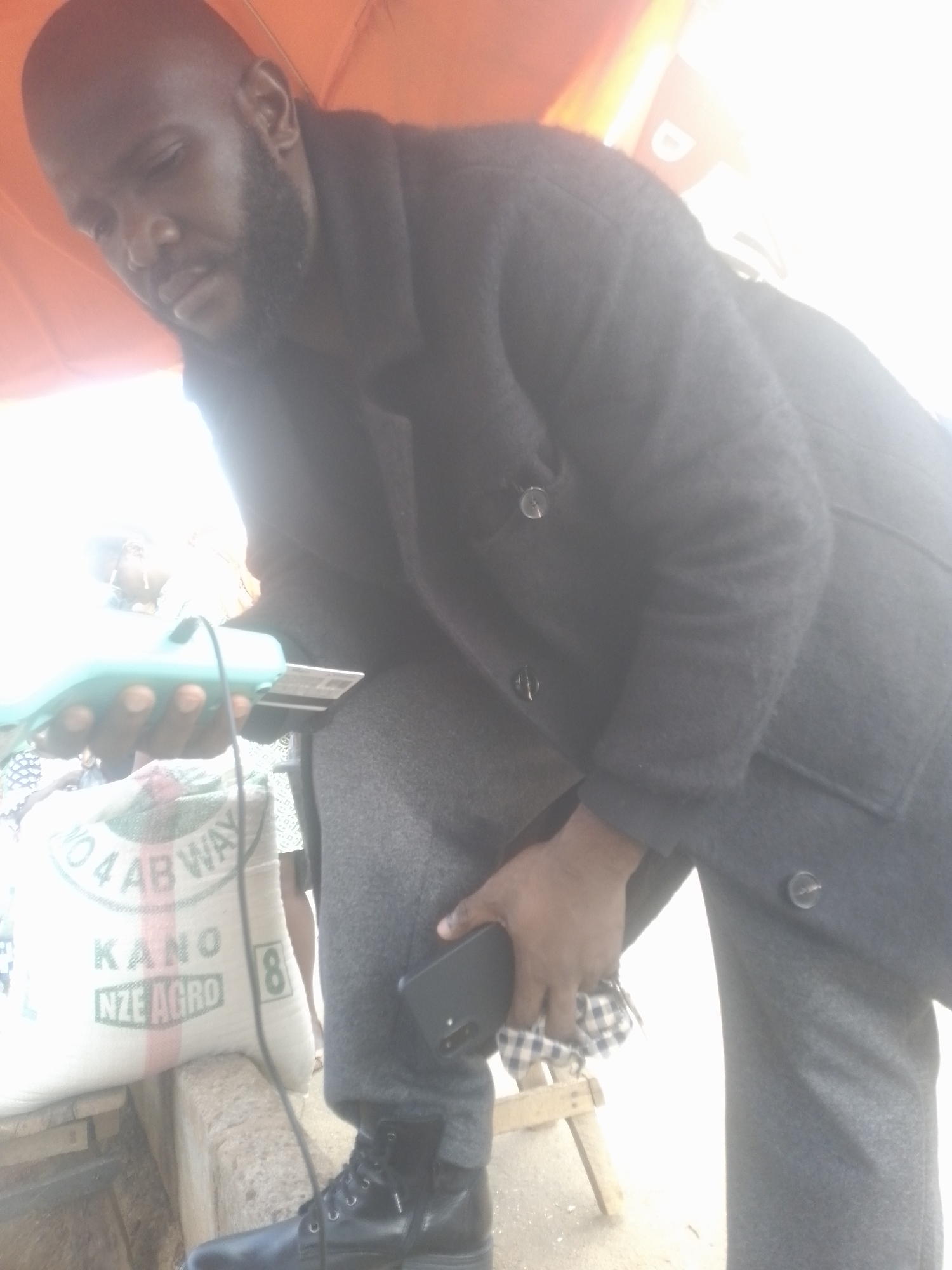 A man with a shaved head and a beard is shown from the waist down, wearing a dark coat and grey pants. He is holding a teal point-of-sale device in his left hand, with a credit card inserted. In his right hand, he is holding a black smartphone and a checkered cloth. He is standing next to a large white sack with green lettering that reads "NO. 4 AB WAY KANO NZE AGRO". The background is blurry and bright, suggesting an outdoor setting with strong sunlight. The image appears to have been taken in an urban environment, possibly a market or street vendor stall.