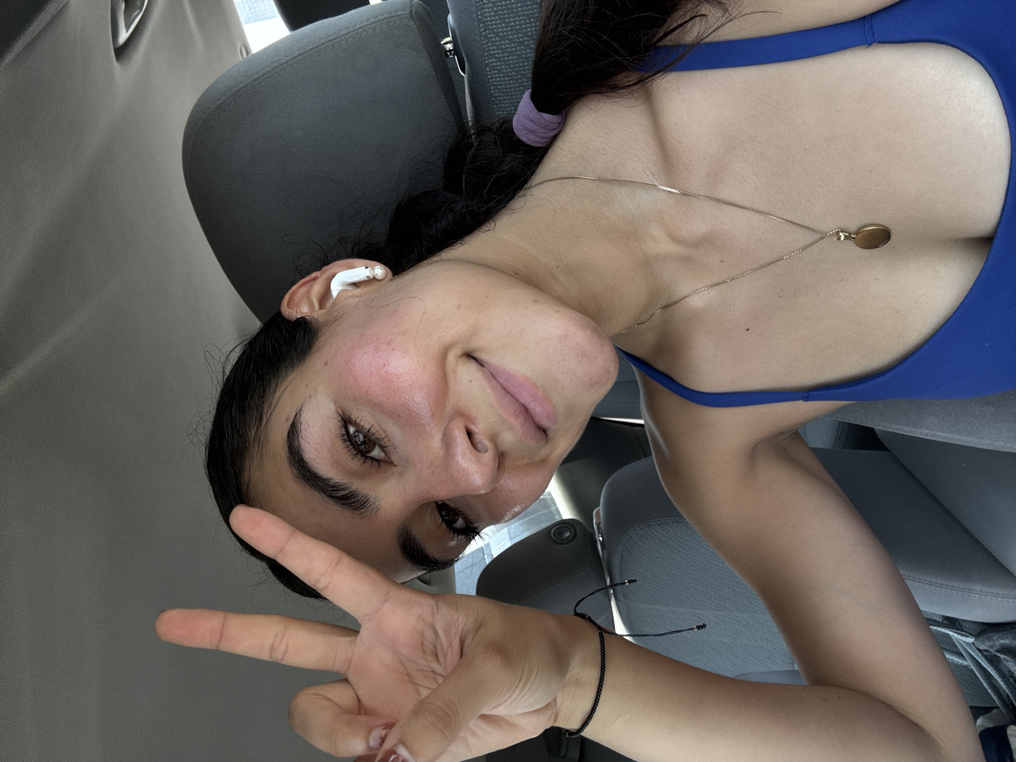 A woman is in the driver's seat of a car. She has dark hair in a braid held by a purple band and is wearing a blue top with a gold necklace. She has an earbud in her left ear and is holding up a peace sign with her right hand. She is wearing a thin black bracelet on her right wrist. The interior of the car is gray. There is no information in the image to identify its location within Puebla, Mexico.