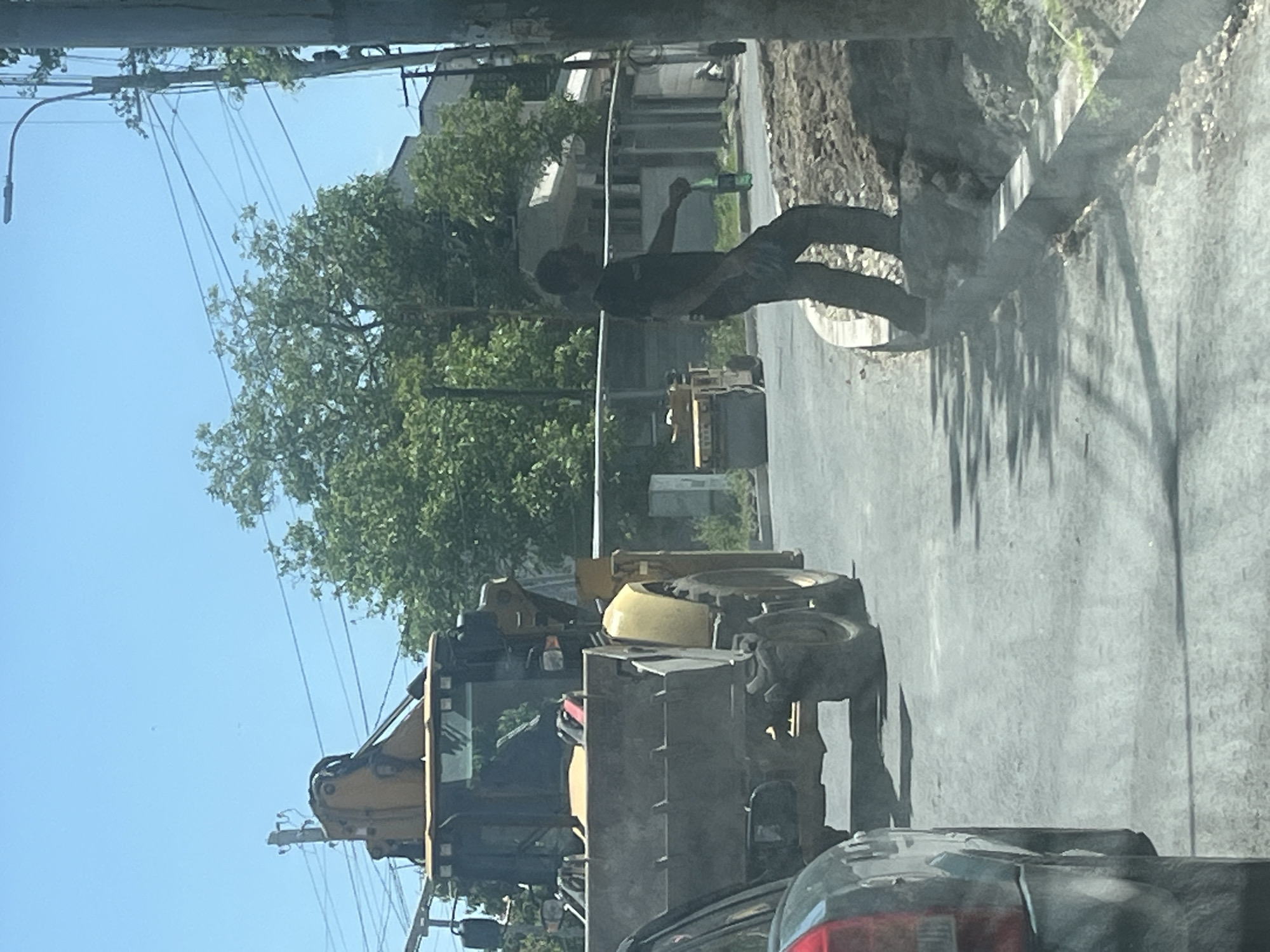 This image depicts an outdoor urban scene, likely in Yerevan, Armenia, characterized by construction or roadwork activity during a bright, sunny day.
The primary subjects include a man, a yellow excavator, and a partially visible dark-colored car. In the mid-ground, a man wearing a dark t-shirt and pants stands on the edge of a paved road next to an area of excavated earth. He is holding a green bottle to his mouth, possibly drinking. His body is slightly turned, and he casts a distinct shadow on the ground behind him.
To the left of the man, a large yellow excavator or backhoe loader is positioned, with its front bucket lowered. The machine appears to be stationary, either parked or in a paused state of operation on the paved road. Only the front section, part of the cab, and a large tire are visible.
In the immediate foreground, the rear portion of a dark car, showing its taillight and bumper, is visible on the right side of the frame, suggesting the photo was taken from inside another vehicle.
The background features dense green trees with heavy foliage under a clear blue sky. Utility poles with multiple overhead power lines are prominent in the upper left section of the image. Behind the construction area and trees, some light-colored buildings can be seen. The ground transitions from a smooth, paved surface to rough, disturbed earth, indicating ongoing work. The time of day is clearly daytime, evidenced by the bright sunlight and strong shadows. There is no discernible text visible in the image.