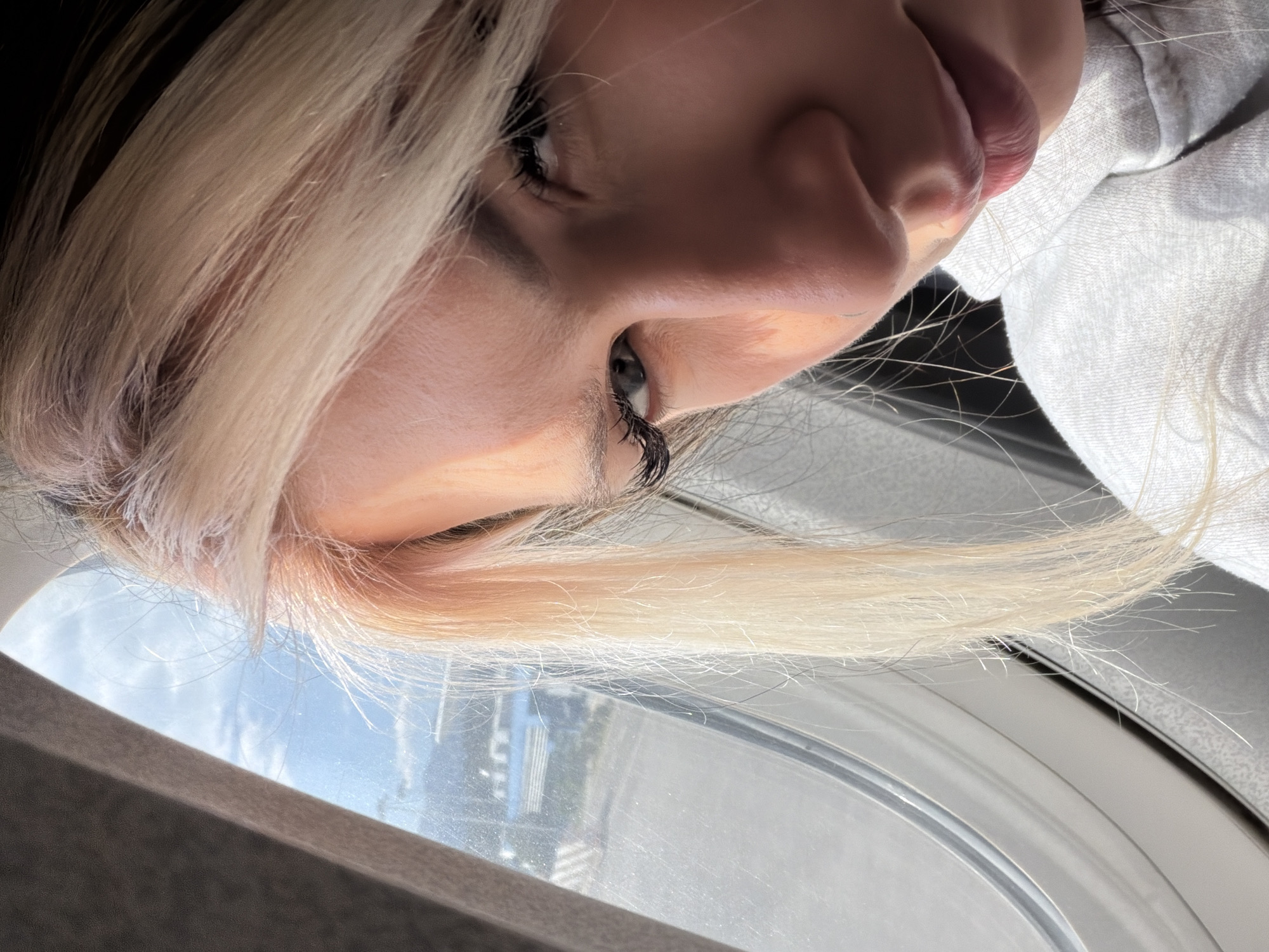 The image captures a close-up, slightly angled view of a person looking out of a window. The main subject is a woman with long, straight, blonde hair, partially obscuring her face. Her left eye is prominently featured, with distinct dark eyelashes and what appears to be a greyish-blue iris. Her nose and lips are also visible.
She is wearing a light-colored garment, possibly a white or light grey shirt or hoodie, visible on the right side of the frame. The window she is looking through has a distinct frame, suggesting it could be an airplane or vehicle window. Outside the window, a bright sky with some clouds is visible, along with distant, blurry shapes that could be buildings or a landscape, indicative of daylight. The overall lighting is bright and natural, casting a soft glow on her features and hair.
The woman appears to be calmly observing the view outside, with no strong emotions discernible from the visible parts of her face. The image is taken at an angle, giving a contemplative or introspective mood. No text is visible in the image. The scene is set in Bogišići, Montenegro.