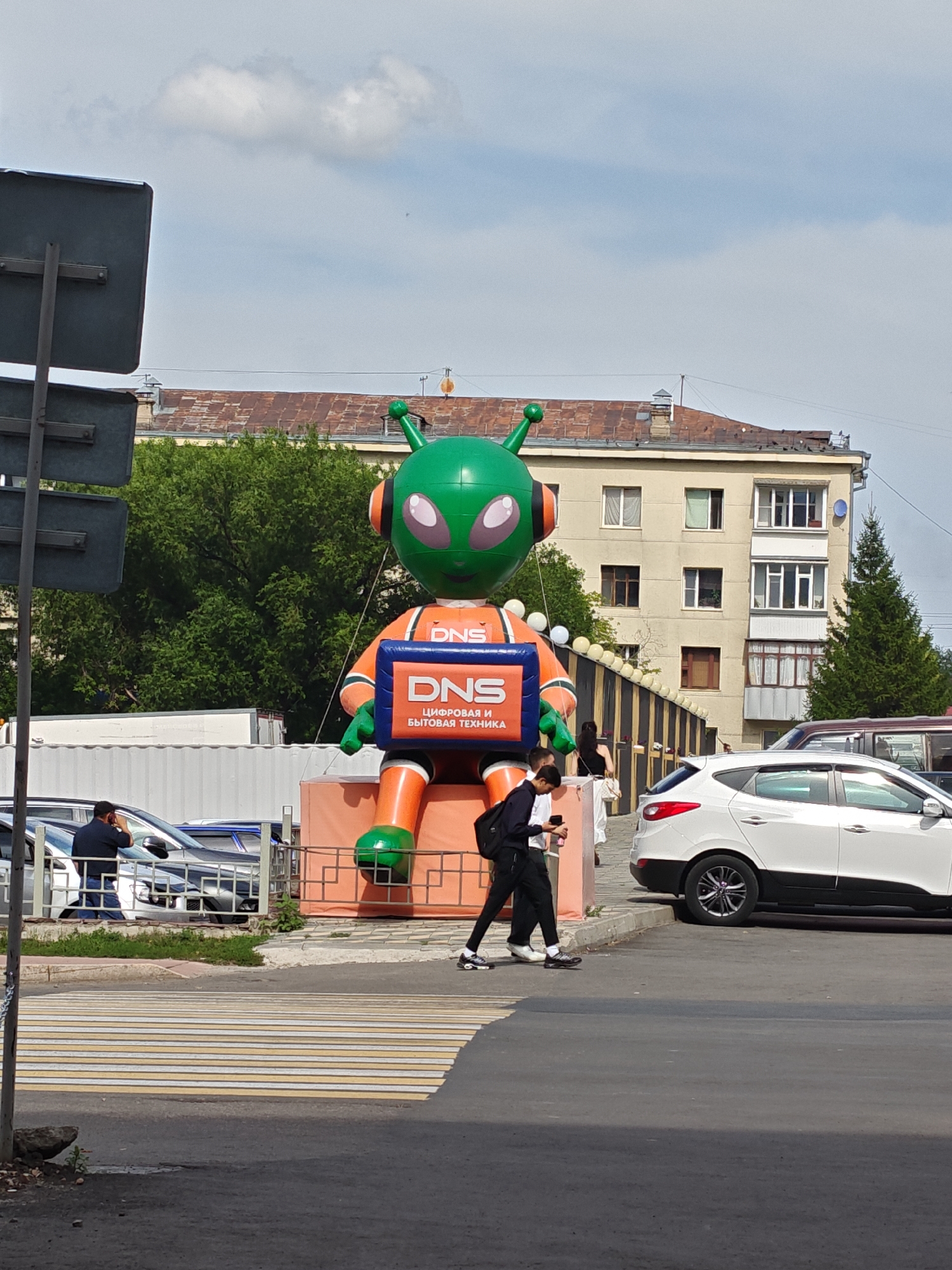 An inflatable alien mascot in orange and green, with a blue box on its lap that reads "DNS" and "ЦИФРОВАЯ И БЫТОВАЯ ТЕХНИКА" (Digital and Home Appliances), is prominently displayed on a raised orange platform. Two individuals are walking on a crosswalk in the foreground. A white car is parked to the right of the scene, and several other vehicles are visible in the background. Behind the inflatable, a building with multiple windows and a tiled roof is visible, along with lush green trees and a cloudy sky. The scene appears to be a street or public area in Petropavl, Kazakhstan.