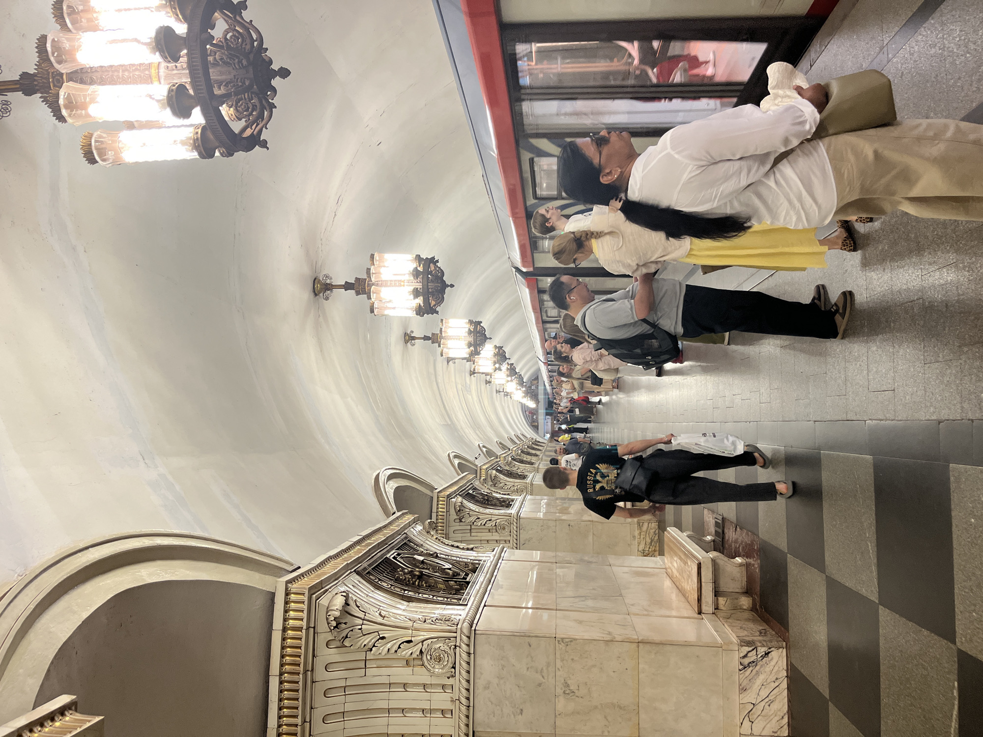 This image depicts a metro station platform in Moscow, Russia, likely part of the Moscow Metro system, known for its ornate stations. The station features a high, arched ceiling with decorative elements and ornate chandeliers. The platform is lined with marble walls adorned with intricate carvings and architectural details. A train is stopped at the platform, with passengers waiting to board or disembark.
In the foreground, a man wearing a black t-shirt with a "RUSSIA" emblem and a black bag slung across his body is walking away from the camera towards the left. He is wearing dark trousers and flip-flops. To his right, a group of people are standing on the platform, facing the train. Among them is a woman wearing a white shirt and beige trousers, holding a light-colored hat in her right hand and a bag over her shoulder. Next to her, a person with blonde hair is wearing a light-colored top and a yellow skirt. A man in a grey t-shirt and black trousers is also visible, wearing glasses and carrying a black backpack.
The platform floor is a checkered pattern of dark and light squares. Benches line the wall on the left side of the image. The train has a red stripe above its windows, and the windows reveal silhouettes of people inside. The overall atmosphere suggests a bustling public transportation hub.