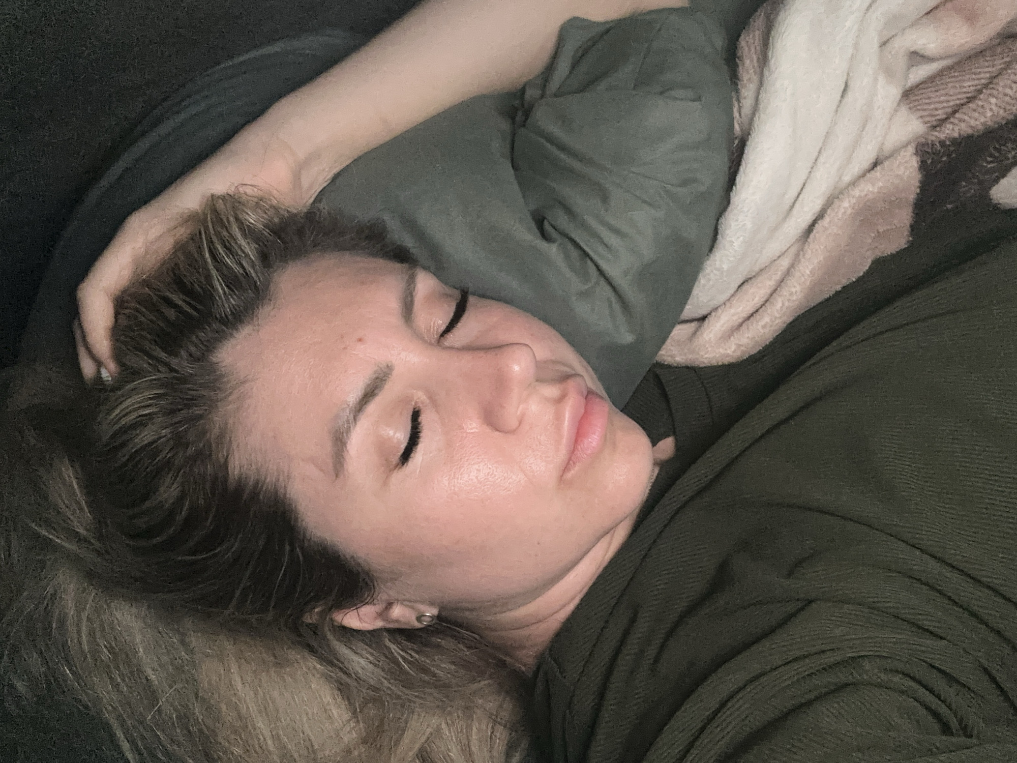A close-up, overhead shot shows a person sleeping on their side. The person's face is partially obscured by a green pillow and a blanket. Their eyes are closed, and their right hand is resting on their head. The person is wearing a dark green, ribbed shirt. The background is dark and out of focus. The scene appears to be indoors, possibly in a bedroom. There is no indication of the location being in Novoaltaysk, Russia.