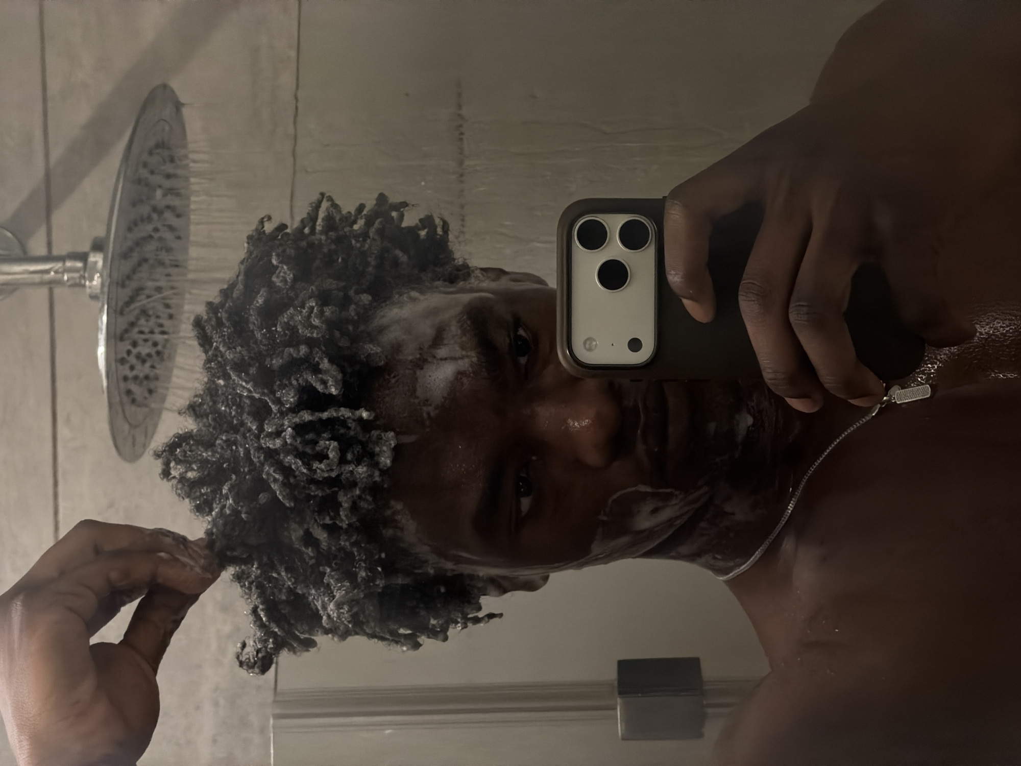 A dark-skinned person is taking a selfie in the shower. The person has short, curly hair that appears to be wet and lathered with soap. A showerhead is visible in the background, with water flowing. The person is holding a phone with three camera lenses, likely an iPhone, in their right hand. The person is wearing a silver chain around their neck. The environment is a bathroom. No information relating to London or the UK is visible in the picture.
