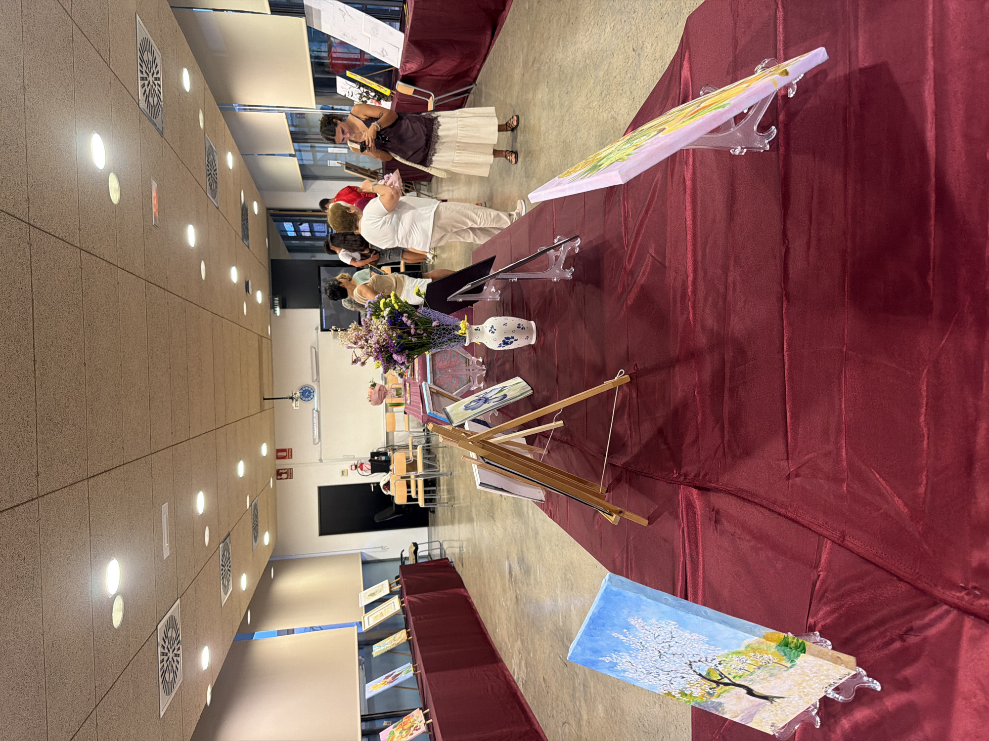 The image depicts an indoor art exhibition, likely in Elche/elx, Spain, given the context. The scene features several tables covered with dark red tablecloths, displaying various paintings on easels and stands. In the foreground, a painting of a tree with white blossoms against a blue sky is prominently displayed. Further back, other paintings are visible, some featuring floral subjects. Several people are present, observing the artwork and interacting with each other. In the middle ground, a woman in a white top and light-colored pants stands near a table, and a woman in a dark top and long skirt is looking at her phone. The environment appears to be a hall or gallery space with a tiled floor and a suspended ceiling with lighting and air vents. Some chairs and other display materials are also visible in the background.