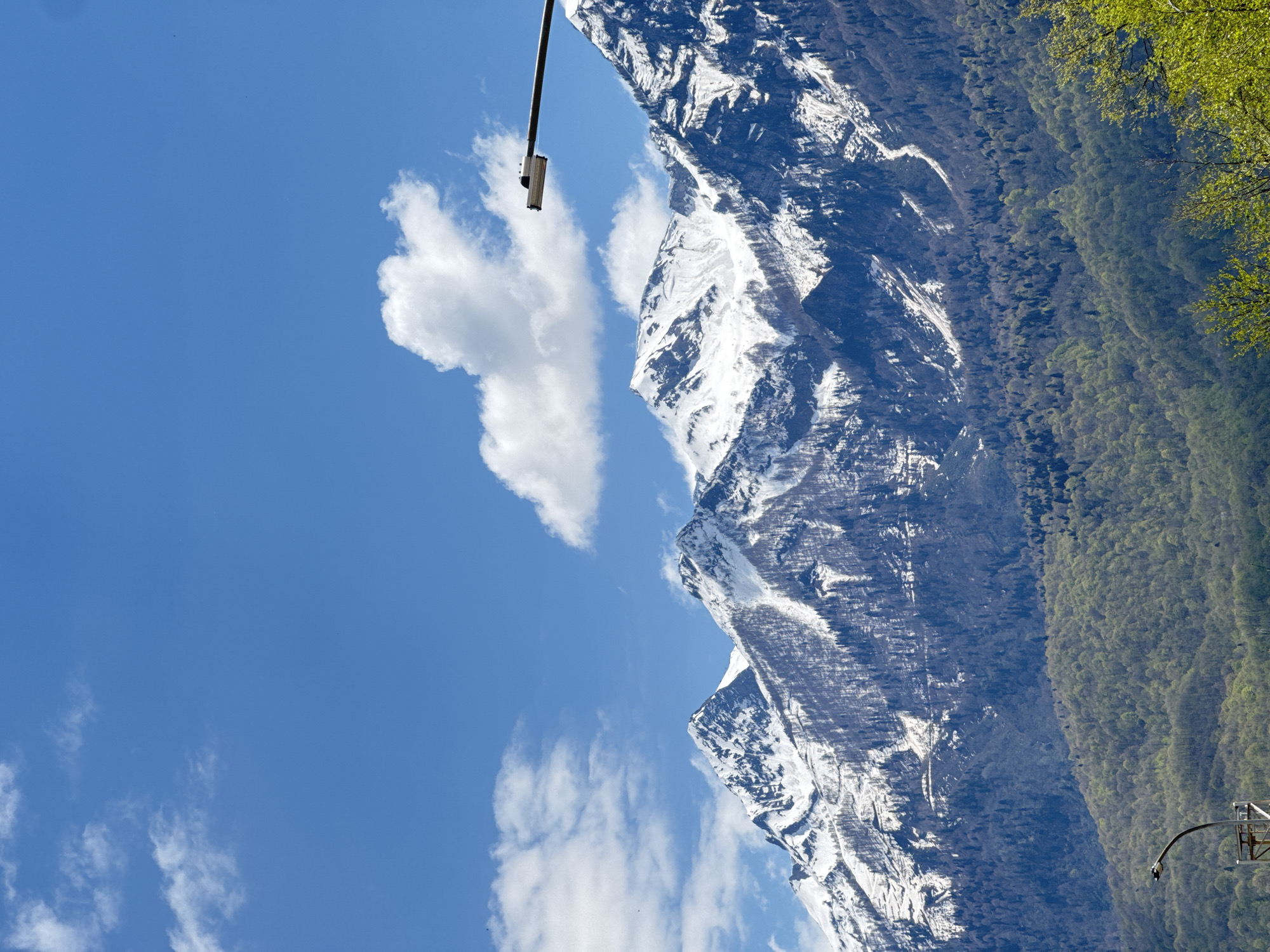 The image shows a mountain range partially covered in snow, with a clear blue sky dotted with white clouds. A dense forest flanks the base of the mountains. A pole with a light fixture is visible in the top left corner, and another pole with a camera-like device in the bottom right corner. The location is likely Krasnaya Polyana, Russia, based on the mountainous terrain and forest density.