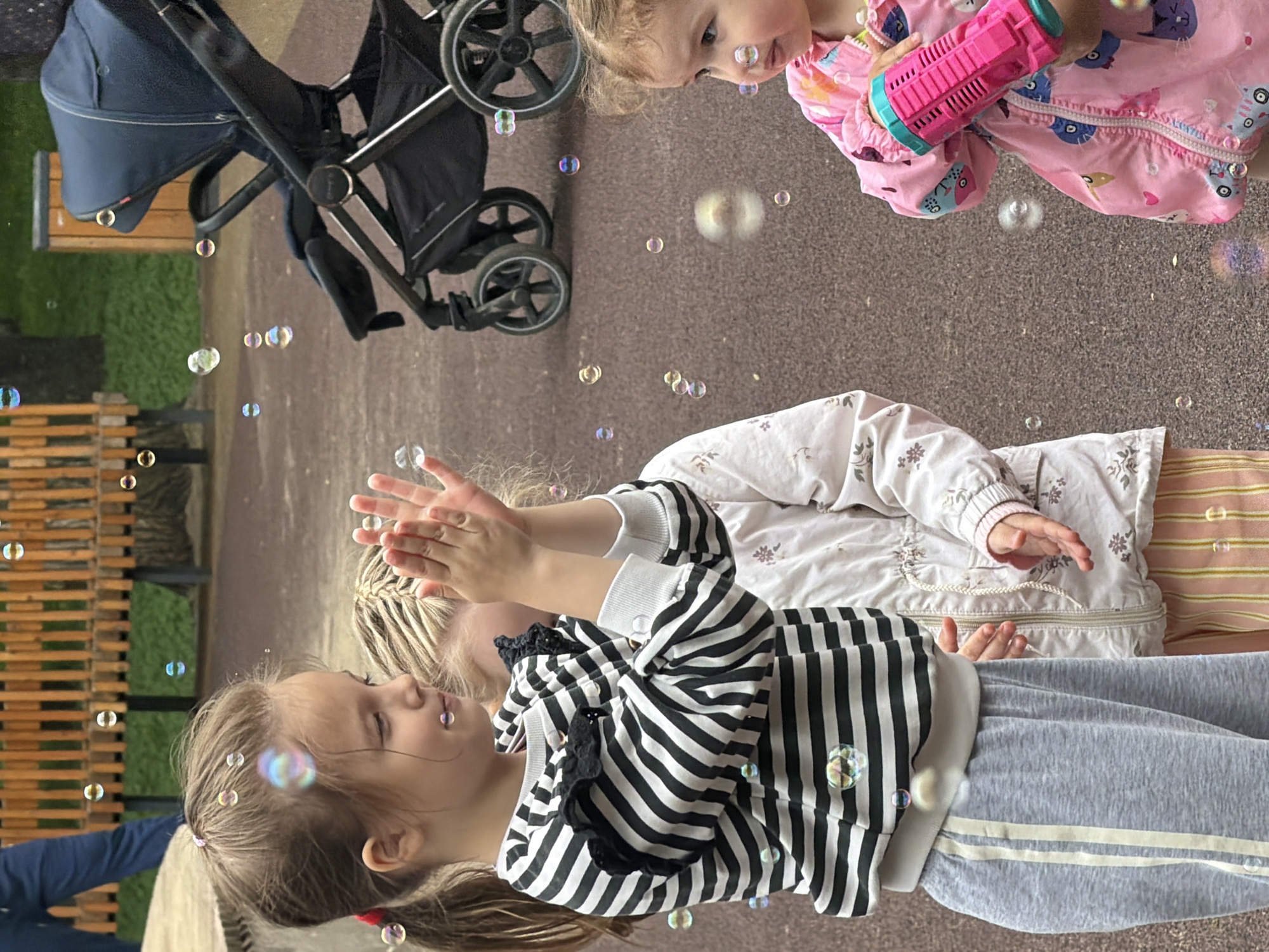 Three young girls are outdoors, playing with bubbles. One girl, wearing a pink patterned jacket, holds a pink bubble-making device. Another girl, dressed in a white floral jacket, reaches towards bubbles with her hands. A third girl, wearing a black and white striped shirt and grey pants, also interacts with the bubbles. A stroller is parked nearby. The setting seems to be a park or recreational area, based on the presence of benches and greenery. There is no indication from the image of the specific city or location in Russia.