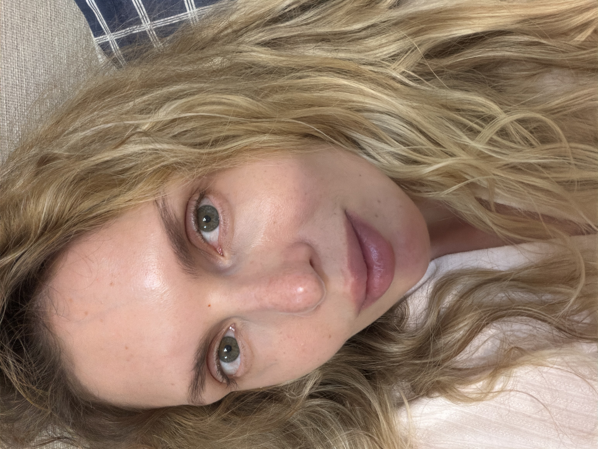 A close-up, angled view of a woman with long, wavy blonde hair. Her head is tilted, and she is looking directly at the camera with slightly widened, green eyes. The woman has fair skin, a small nose, and pink lips. She appears to be lying down, as indicated by the position of her head and the presence of a white fabric or possibly a pillow beneath her hair. In the upper left corner of the image, there is a glimpse of a blue and white checkered pattern, which could be part of a pillow or blanket. There are no indicators suggesting that the image was taken in the city of Budva, Montenegro.