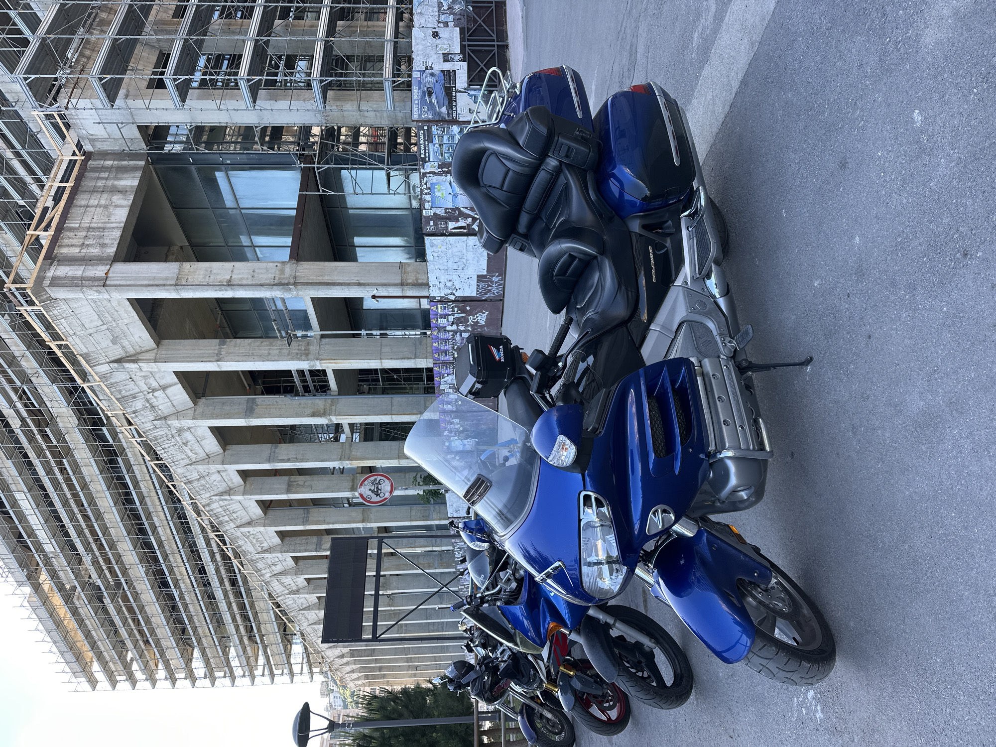 The image shows a row of parked motorcycles on a paved surface, likely a street. Dominating the foreground is a large, blue touring motorcycle with a tall windscreen and prominent fairing. Behind it are several other motorcycles, decreasing in visibility due to perspective.
In the background is a large, unfinished building under construction. The building's structure is exposed, showing concrete framework and scaffolding. To the right of the building, posters and graffiti cover a concrete wall. A circular sign with a motorcycle symbol crossed out is affixed to the building.
The image was taken in Budva, Montenegro, as evidenced by the distinctive architectural style of the unfinished building and the prevalence of motorcycles often found in urban European settings. The perspective and positioning of the motorcycles suggest they are parked along a street adjacent to the construction site.