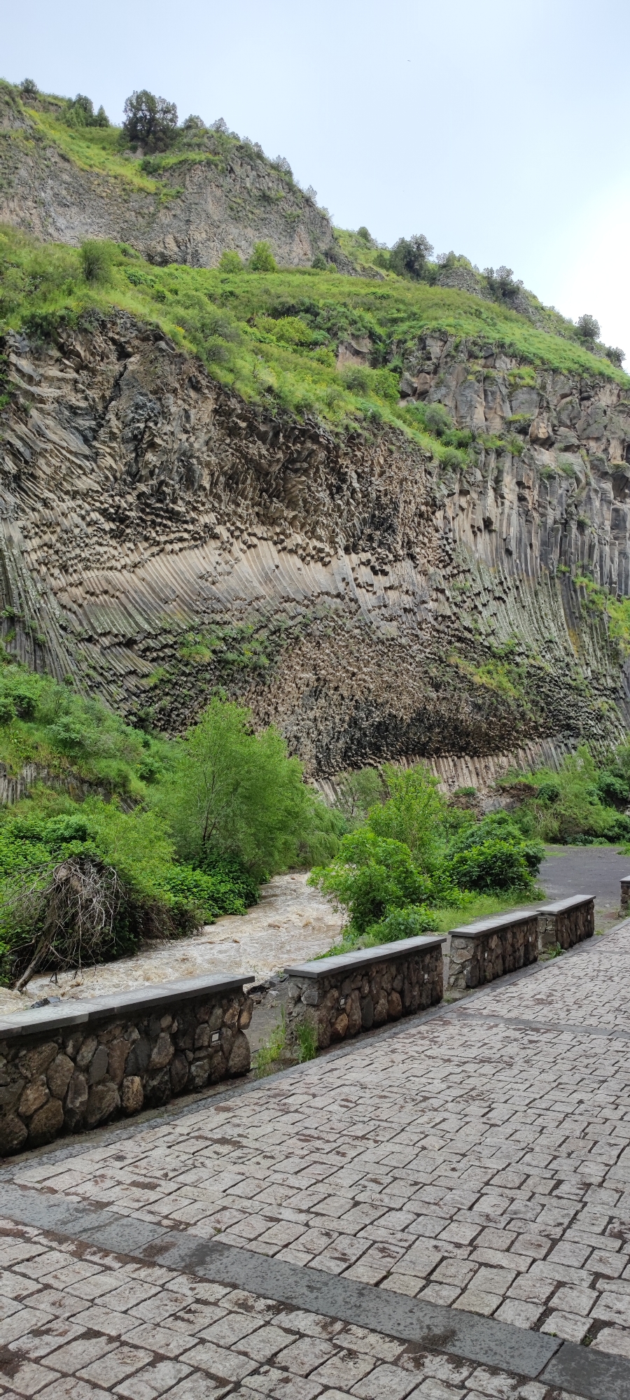 The image depicts a view of the Symphony of Stones, a basalt column formation, within the city of Garni, Armenia. The scene features a river with muddy-colored water flowing alongside a stone-paved walkway. A low stone wall separates the walkway from the river. Green vegetation is visible along the riverbank and on the hillside above the rock formation. The basalt columns display a honeycomb-like pattern in some sections, and vertical columns in others. The sky appears overcast.