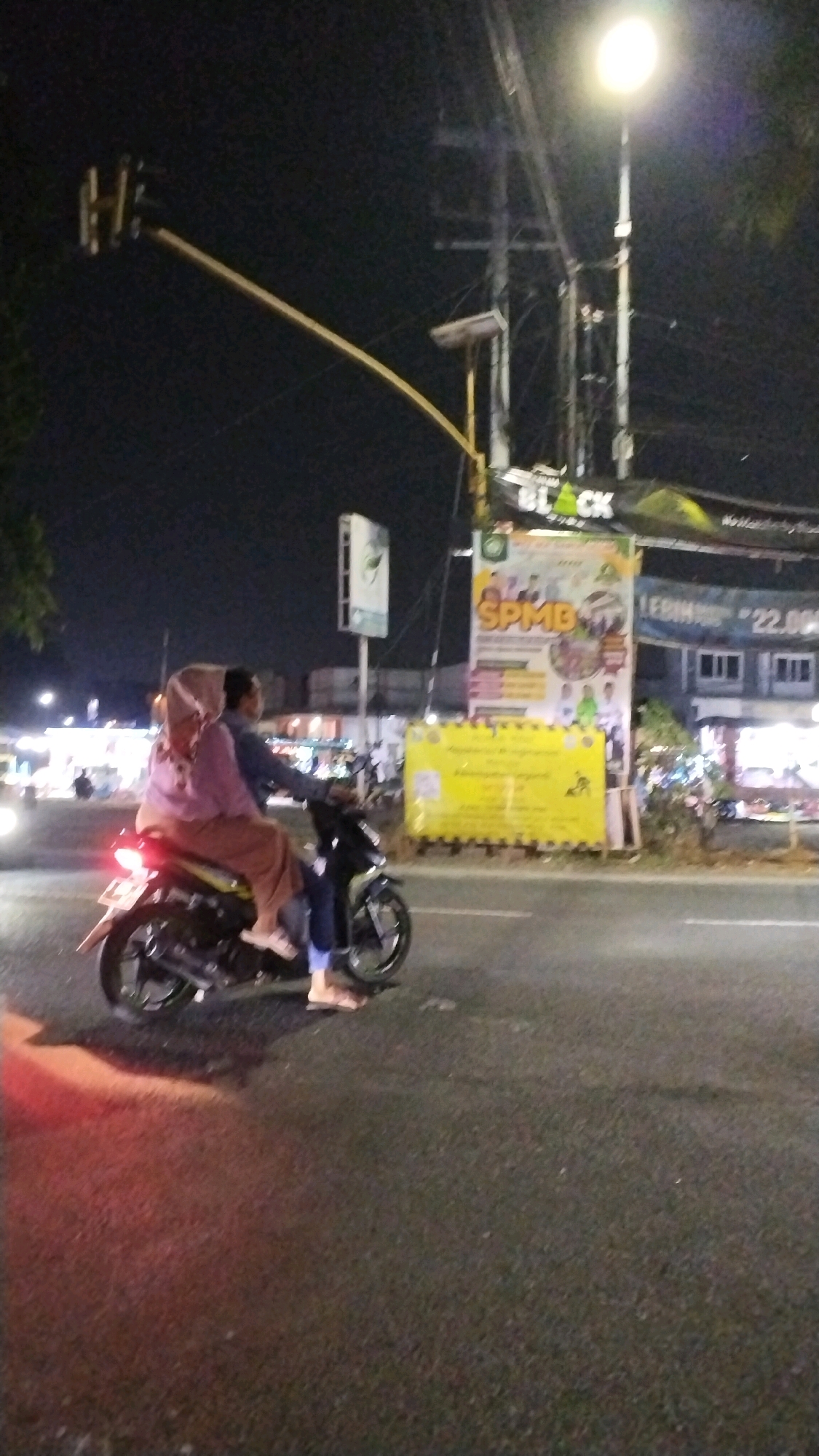 A slightly blurry, night-time, eye-level shot captures a motorcycle with two occupants on a paved road in Canggu, Indonesia. The rider, a male with dark hair, wears a blue long-sleeved shirt and dark pants. The passenger, a female, is wearing a light purple hijab and a brown skirt, and is seated behind the rider. Both are facing to the right. The motorcycle is a dark color and appears to be in motion, with its rear brake light illuminated. In the background, a street light illuminates the scene from the top right, casting a bright glow. A traffic light hangs from a pole on the left side of the image. Two billboards are visible in the background; one is yellow with text, and another, directly behind it, has the word "SPMB" prominently displayed in large orange letters. Buildings and other indistinct structures line the street. The overall lighting suggests it is nighttime.