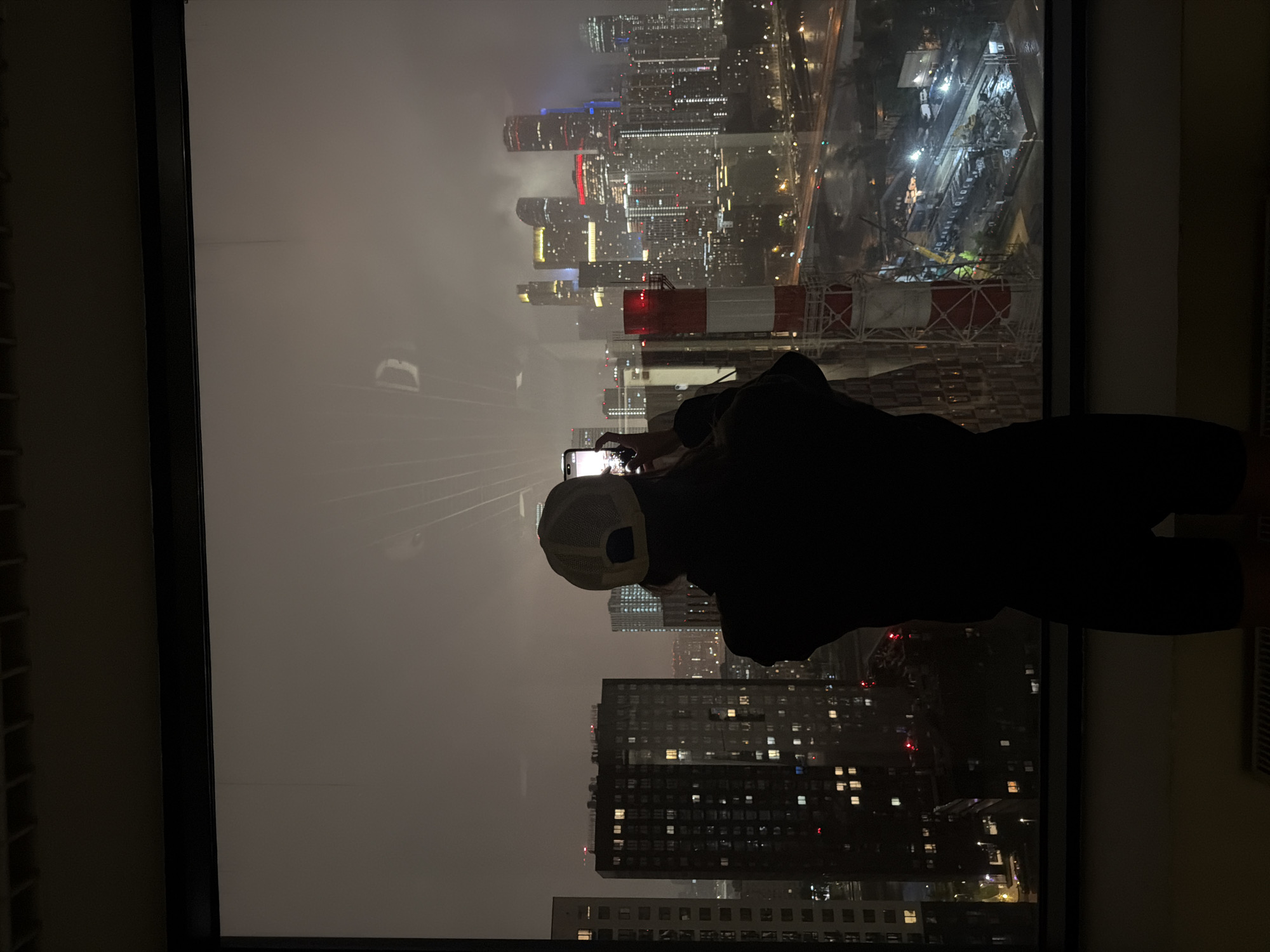 A person, facing away from the camera, stands at a large window, holding a smartphone to take a photo of the cityscape. They are wearing a dark jacket and a light-colored baseball cap. Through the window, a dense urban environment is visible at night, characterized by numerous tall buildings with illuminated windows. In the foreground, a tall, red and white striped industrial smokestack or tower dominates the view. The city lights are bright, reflecting off a wet surface, suggesting it may be raining. The overall atmosphere is dark and moody, with the lights of the city piercing through the hazy, overcast sky. The location is the city of Khimki, Russia.