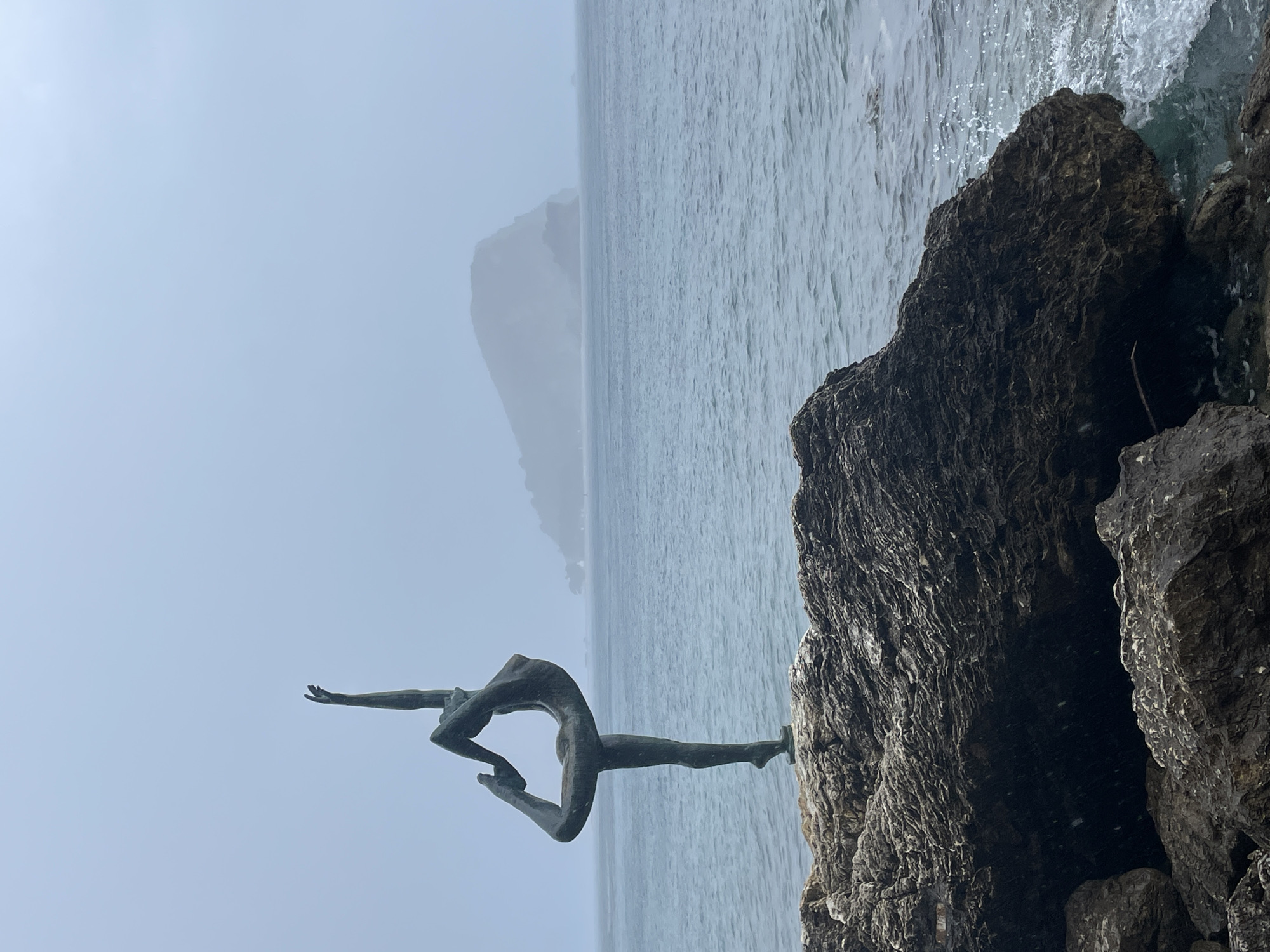 The image depicts the "Dancer from Budva" (Ballerina) statue in Budva, Montenegro, located on the rocky coastline near Mogren Beach. The bronze-colored statue of a slender female figure is poised in an arabesque, with one leg extended backward and arms reaching upwards, balanced on the ball of her right foot on a flat surface atop a large, dark, textured rock.
The foreground consists of rugged, dark grey rock formations, some showing signs of erosion and exposure to water. To the right and behind the statue, the Adriatic Sea extends, displaying choppy, dark blue-grey water with visible small waves and sea foam near the rocks. In the distant background, a large, hazy landmass, likely Sveti Nikola Island, is partially obscured by mist or a grey, overcast sky. The sky itself is a uniform light grey, blending into the distant horizon. The overall scene suggests an overcast day with potentially light precipitation, indicated by small white specks in the air, particularly visible in the lower portions of the image. No other individuals or specific objects are visible.