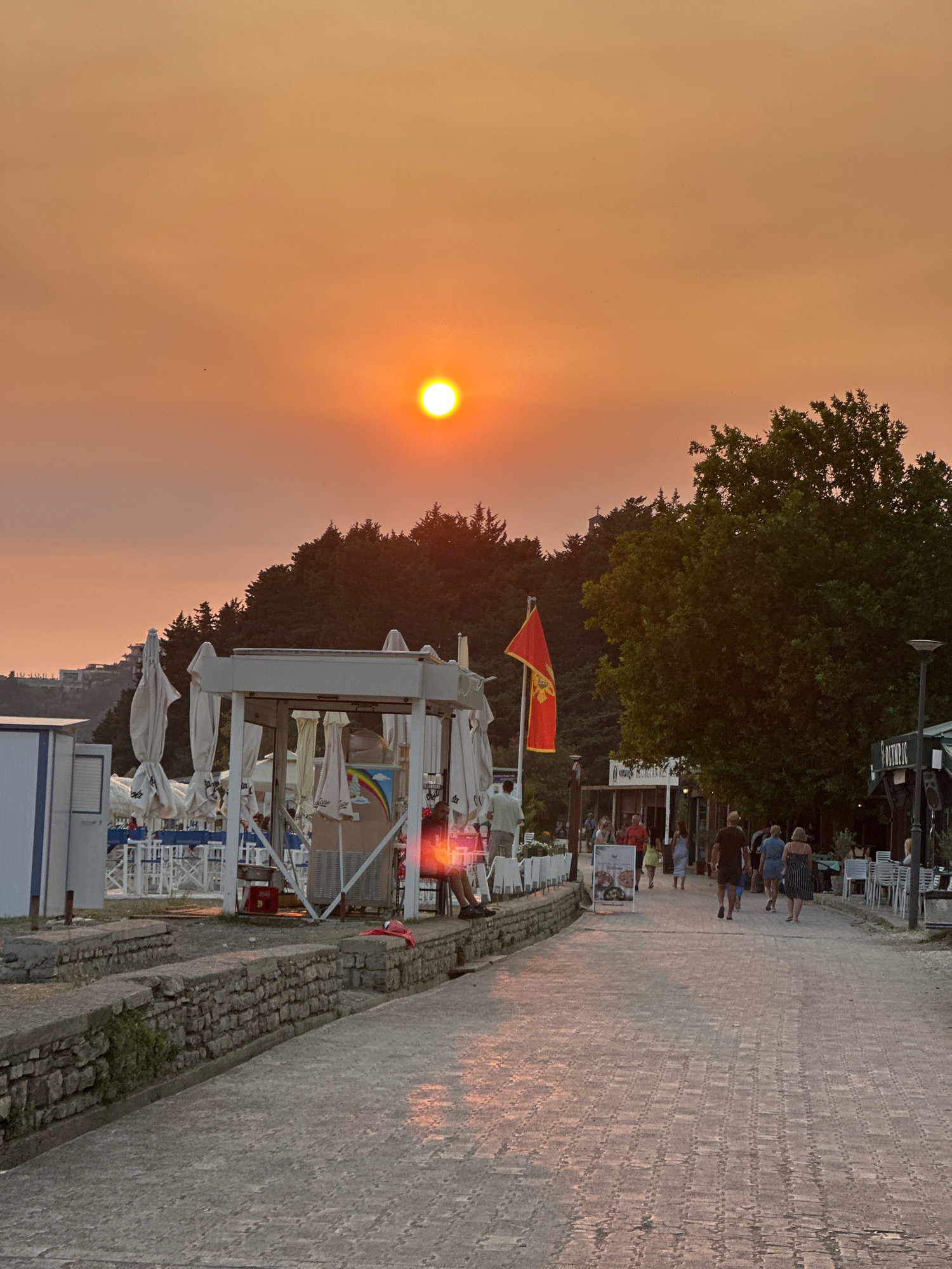 The image depicts a pedestrian pathway in Boreti, Montenegro, during sunset. The sky is a vibrant orange, indicating either sunset or the presence of smoke, with the sun appearing as a bright, diffused disc. On the left side of the pathway, there is a row of white beach umbrellas and seating areas, suggesting a beach or seaside resort. A few people are visible sitting and walking in this area.
Further down the pathway, several small buildings and structures are visible, some appearing to be shops or cafes. A prominent red flag with a yellow emblem, possibly the flag of Montenegro, is mounted on a flagpole. People are walking along the paved pathway, some in small groups, and others individually. The pathway is made of light-colored paving stones, which reflect the warm light of the sunset. Dense green trees line the right side of the pathway, forming a backdrop to the scene. In the distance, a hilly landscape with some buildings is visible, partially obscured by trees. The overall atmosphere suggests a late afternoon or early evening at a coastal location.