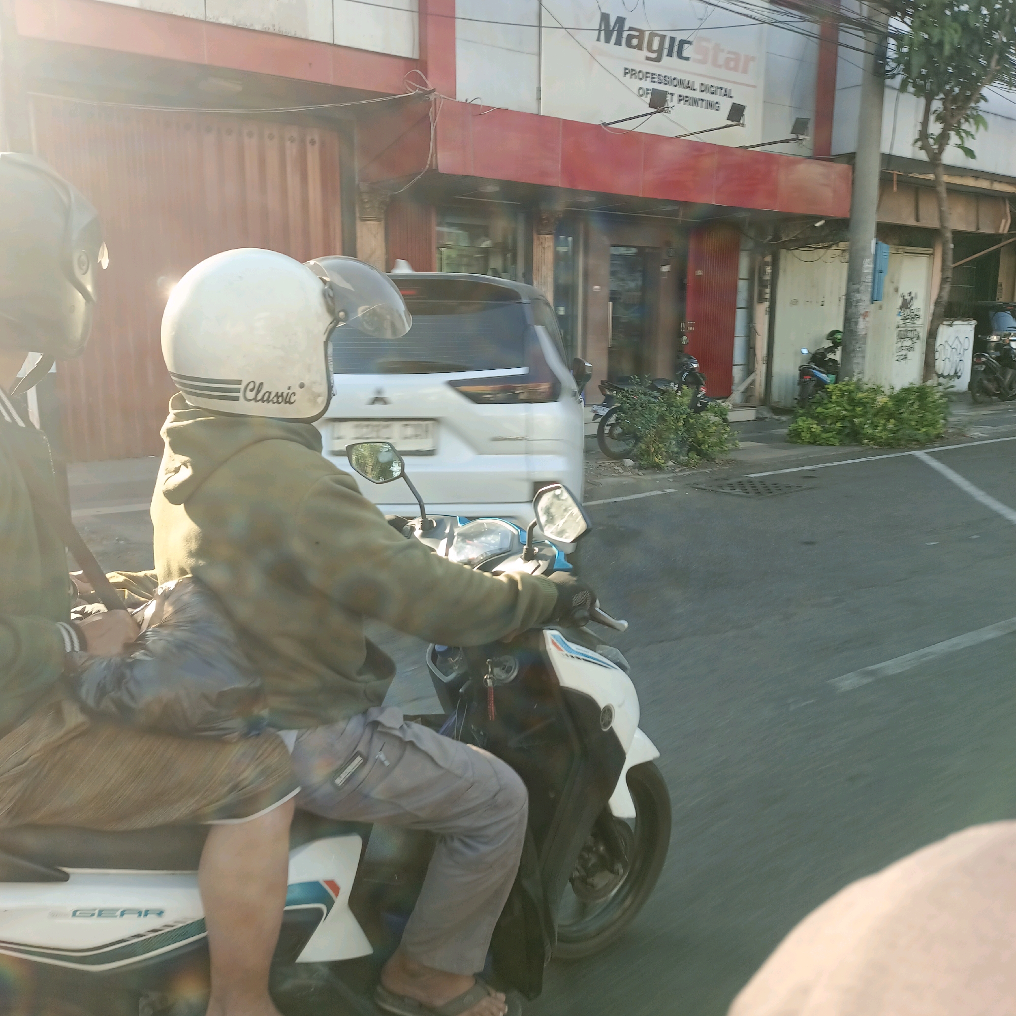 The image depicts a sunny street scene in Surabaya, Indonesia. In the foreground, two individuals are riding a white scooter. The person in the front is wearing a white helmet with the word "Classic" visible, a green hoodie, and grey pants. They are holding the handlebars and appear to be in motion. The second person, behind the driver, is wearing a dark helmet and a green jacket. They are seated behind the driver, with their arms around them, and have a dark bag draped over their lap.
The scooter has a white body with blue and red accents and the word "GEAR" is visible on the side. The scooter has visible mirrors and a license plate on the rear. In the background, a white SUV is partially visible, also in motion, with a license plate that is difficult to read.
Adjacent to the street is a building with a red facade and large red doors. Above the red facade, a white building with the prominent text "MagicStar PROFESSIONAL DIGITAL OFFSET PRINTING" is visible. The street is paved and appears to be a city road, with a white lane marking visible. The lighting suggests it is daytime, with bright sunlight casting lens flares and highlighting parts of the scene. Various parked motorbikes and lush green foliage are also present along the street, indicating a commercial or residential area in Surabaya.