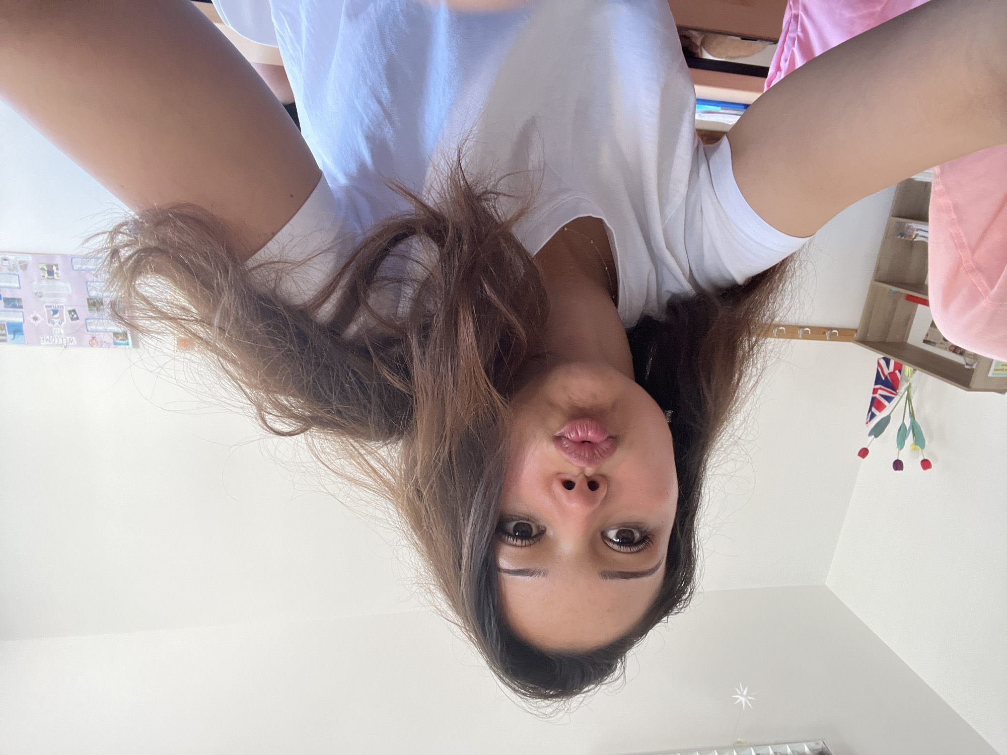 A young woman with long, dark brown hair is taking a selfie in what appears to be a classroom or office environment. She is wearing a white t-shirt and has her lips puckered for a kiss. Behind her, a white wall is visible, with some shelves containing books and decorations, including a Union Jack flag and some colorful flowers. A desk is also partially visible in the foreground. The overall lighting suggests an indoor setting with artificial light from above. The specific location in Zlatograd, Bulgaria is not evident from the visual information in the image.