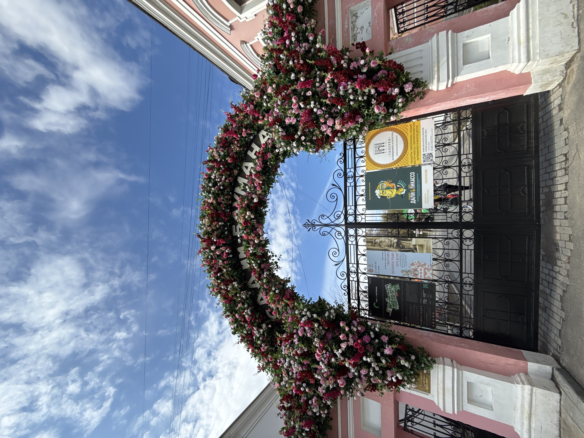 The image captures a unique architectural display in Moscow, Russia. An arched entrance, adorned with an extravagant floral arrangement of red, pink, and white flowers and greenery, spans the gate. On the gate are several posters, including one advertising the gallery of antiquities and culture "Другое Дело" at 15 Staraya Basmannaya street, as well as a Dali & Picasso exhibit. Another poster advertises an exhibition at the Nicholas Museum, “In Faith, Loyalty, Work", from April 25th to May 15th. The architectural style of the building surrounding the gate features a pink facade with white trim, indicating a blend of classic and modern designs. The sky, a clear blue with scattered white clouds, serves as a backdrop to the floral arch. The scene suggests an artistic or cultural event, given the presence of the posters and the ornate floral decorations.