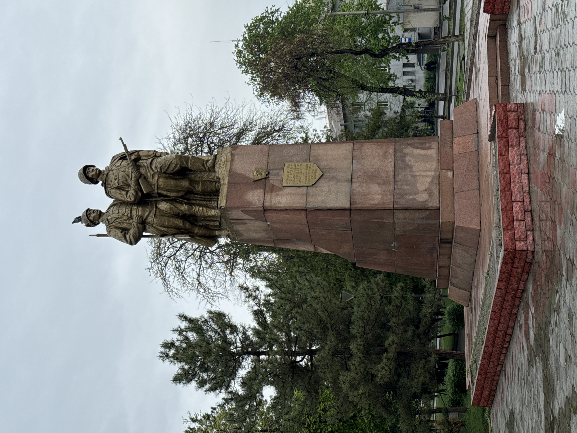 The image displays a large war memorial featuring two soldier statues atop a multi-tiered stone pedestal, set under an overcast sky in what appears to be a public square or park in Bishkek City, Kyrgyzstan.
The central subjects are two male soldier figures, sculpted in bronze or a similar material, standing side-by-side. Both are depicted wearing military uniforms, helmets, and holding rifles, poised as if on watch or in formation. The statues have a weathered, greenish-brown patina. Their faces are solemn, reflecting the commemorative nature of the monument.
The statues rest on a substantial pedestal constructed from reddish-brown stone, possibly granite, arranged in several receding tiers. A bronze-colored plaque, shaped like a shield, is affixed to the front of the pedestal. The inscription, in Cyrillic script, is partially legible and includes phrases such as "ВЕЧНАЯ ПАМЯТЬ" (Eternal Memory), "ГЕРОЯМ" (to heroes), and "ПОГИБШИМ" (who died), along with the year "1941." The bottom portion of the inscription also appears to read "ОТ БЛАГОДАРНЫХ СООТЕЧЕСТВЕННИКОВ" (from grateful compatriots), indicating a dedication to fallen soldiers, likely from World War II. A small, indistinct circular emblem is visible to the left of the plaque.
The monument is surrounded by greenery, including both deciduous trees with early spring foliage or bare branches, and evergreen pine trees. The ground is paved with gray, interlocking blocks, which appear damp, suggesting recent rain. In the background, a multi-story building with visible windows can be seen through the trees. The sky is uniformly gray and cloudy, casting a soft, muted light over the scene. The overall atmosphere is one of solemn remembrance, typical of a war memorial.