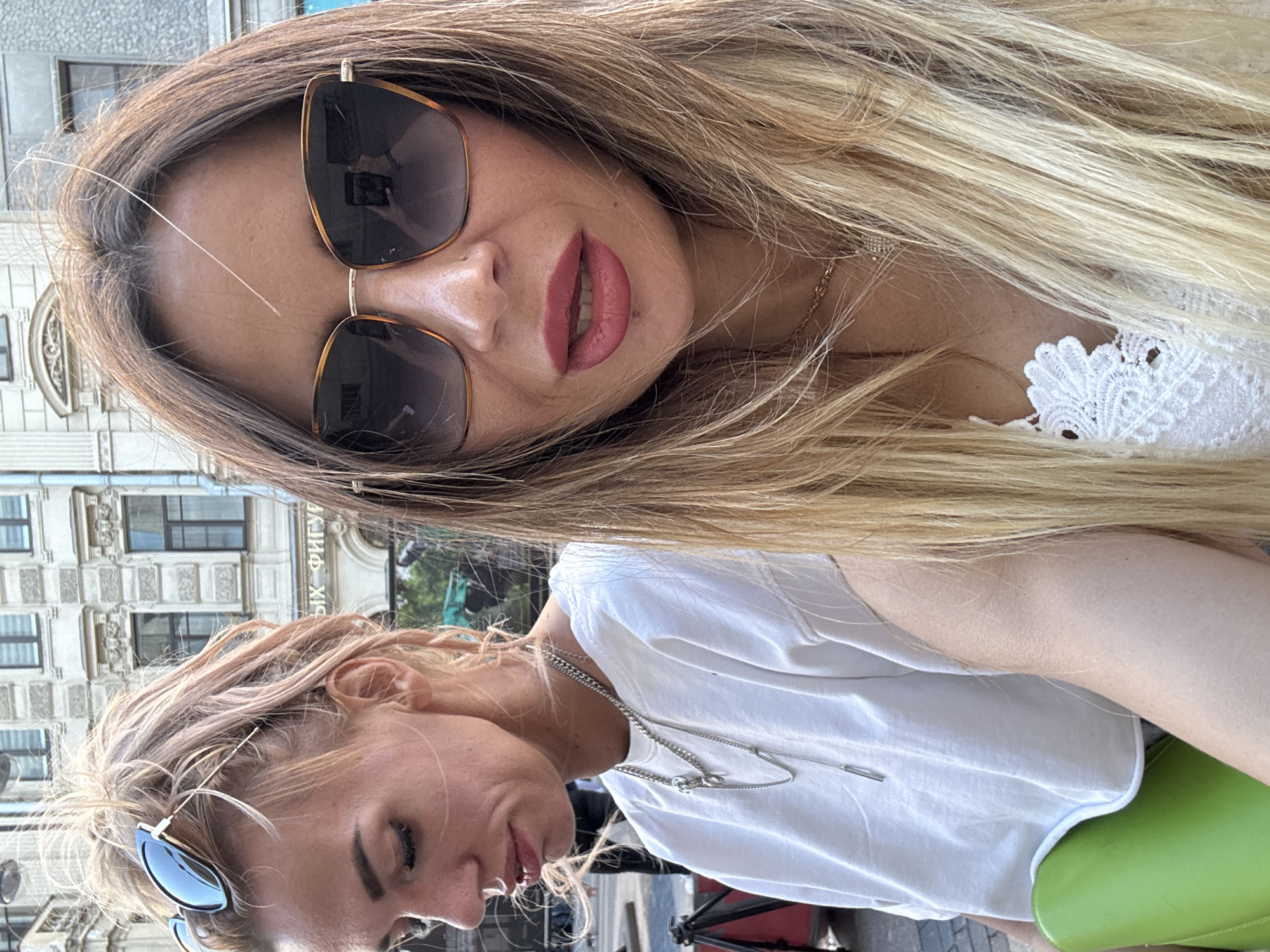 This media file features two women in an outdoor setting, likely a street or cafe in Saint Petersburg, Russia. The image is a selfie, taken at eye level, with the camera tilted slightly.
The woman in the foreground, on the left, has blonde hair and is wearing sunglasses and a white t-shirt with a silver necklace. She is looking down and to the right, with a slight smile.
The woman in the background, on the right, has long blonde hair and is wearing sunglasses and a white top with lace detailing. She is looking towards the camera, with her lips slightly parted and a hint of pink lipstick. Her expression is playful and inviting.
The background shows a building with architectural details that suggest a historic European city. The lighting indicates it is daytime, likely sunny, as indicated by the shadows and the overall brightness of the scene.
The image captures a moment of casual interaction between two friends, enjoying a day out. The mood is light and cheerful, reflecting the warm weather and pleasant company. There is no visible text in the image.