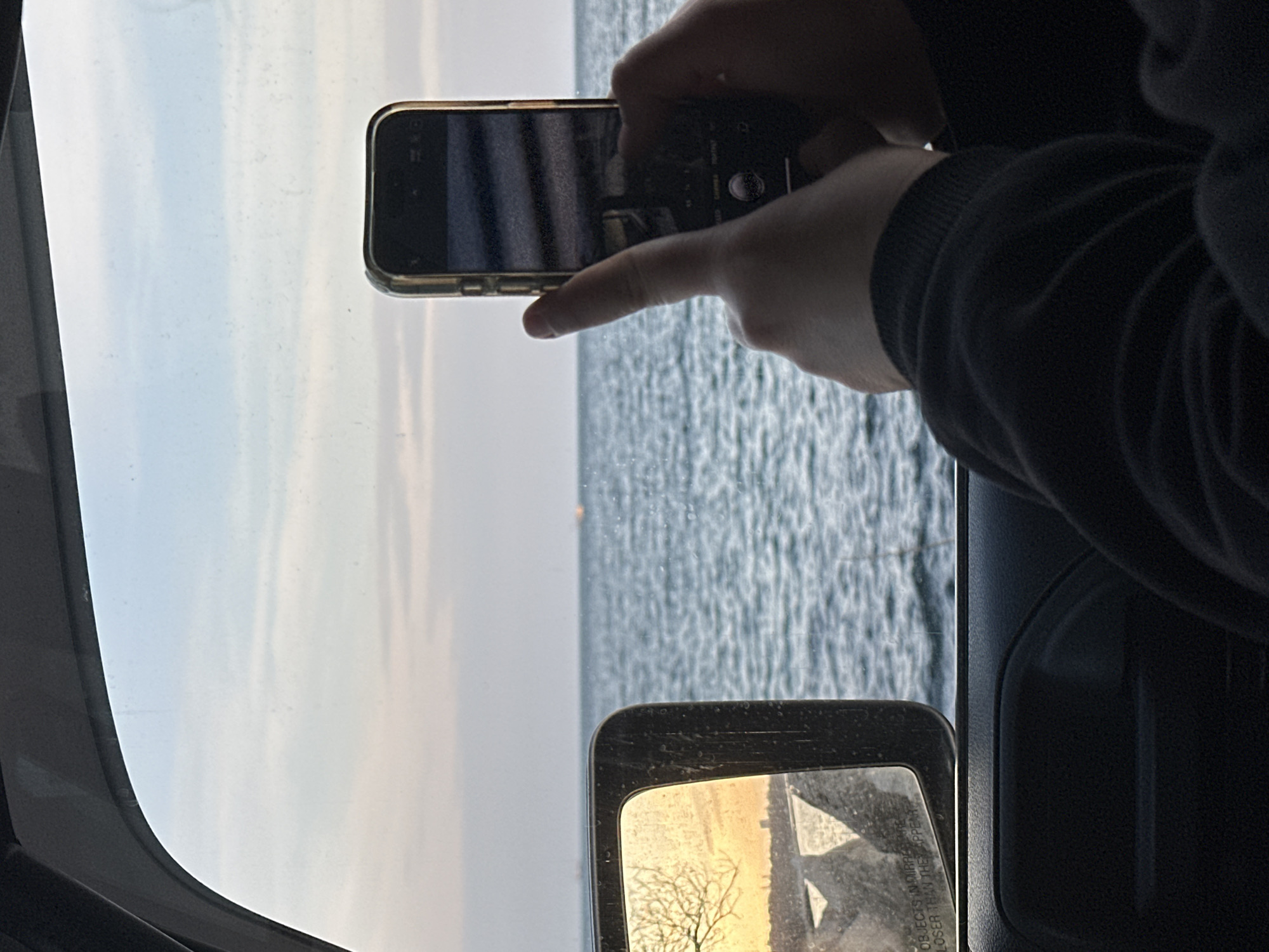 The image shows a person inside a vehicle, possibly a car or truck, taking a photo with a smartphone. The view out of the vehicle's window reveals a body of water, likely the Black Sea, with a light-colored sky. A vehicle side mirror is visible. The reflection in the mirror appears to show a view of trees, possibly indicating a location near land rather than a completely open sea view. This could be in Varna, Bulgaria.